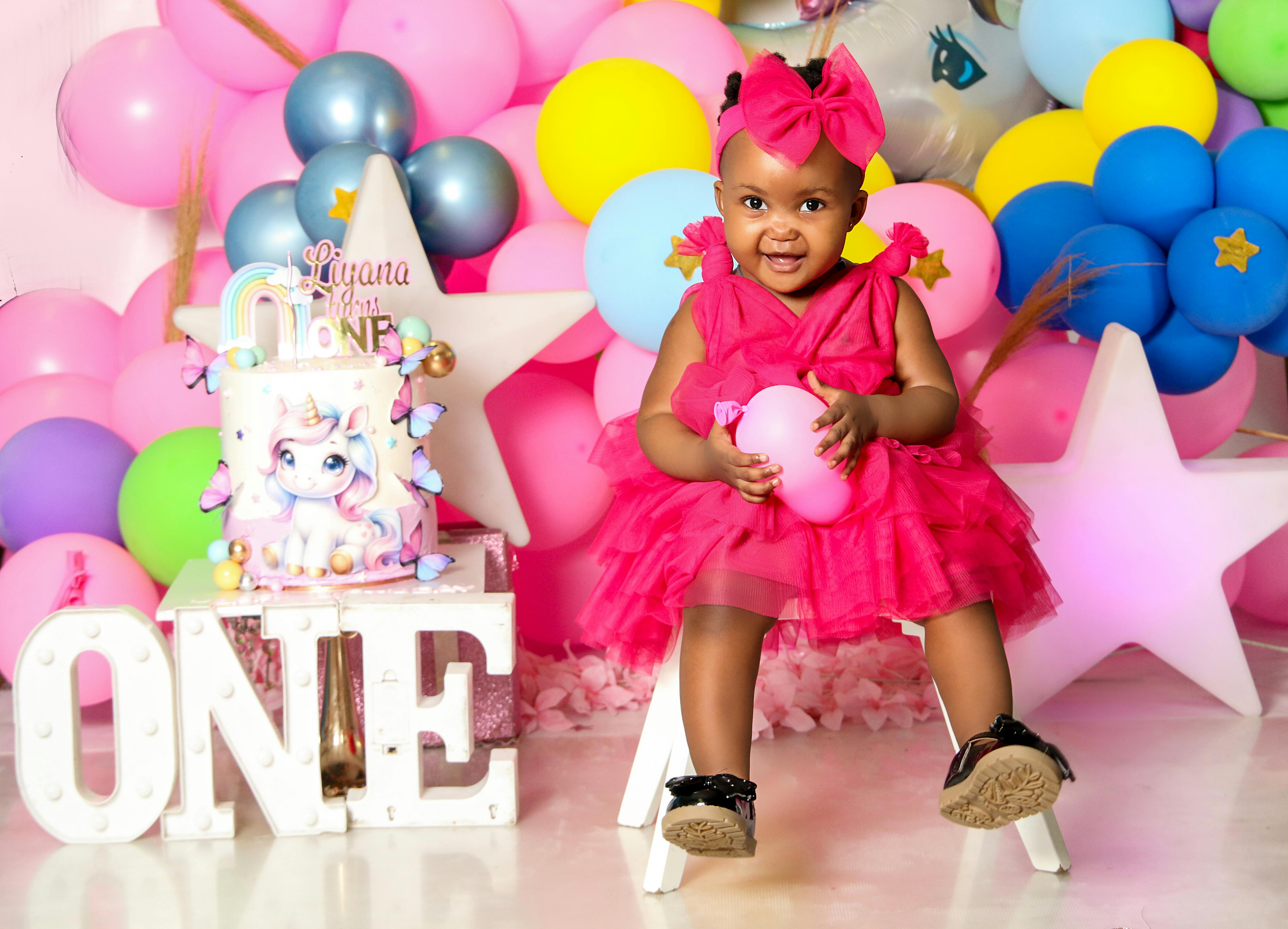 A joyful first birthday party setup with a cute child in a vibrant pink dress surrounded by balloons and a unicorn cake.