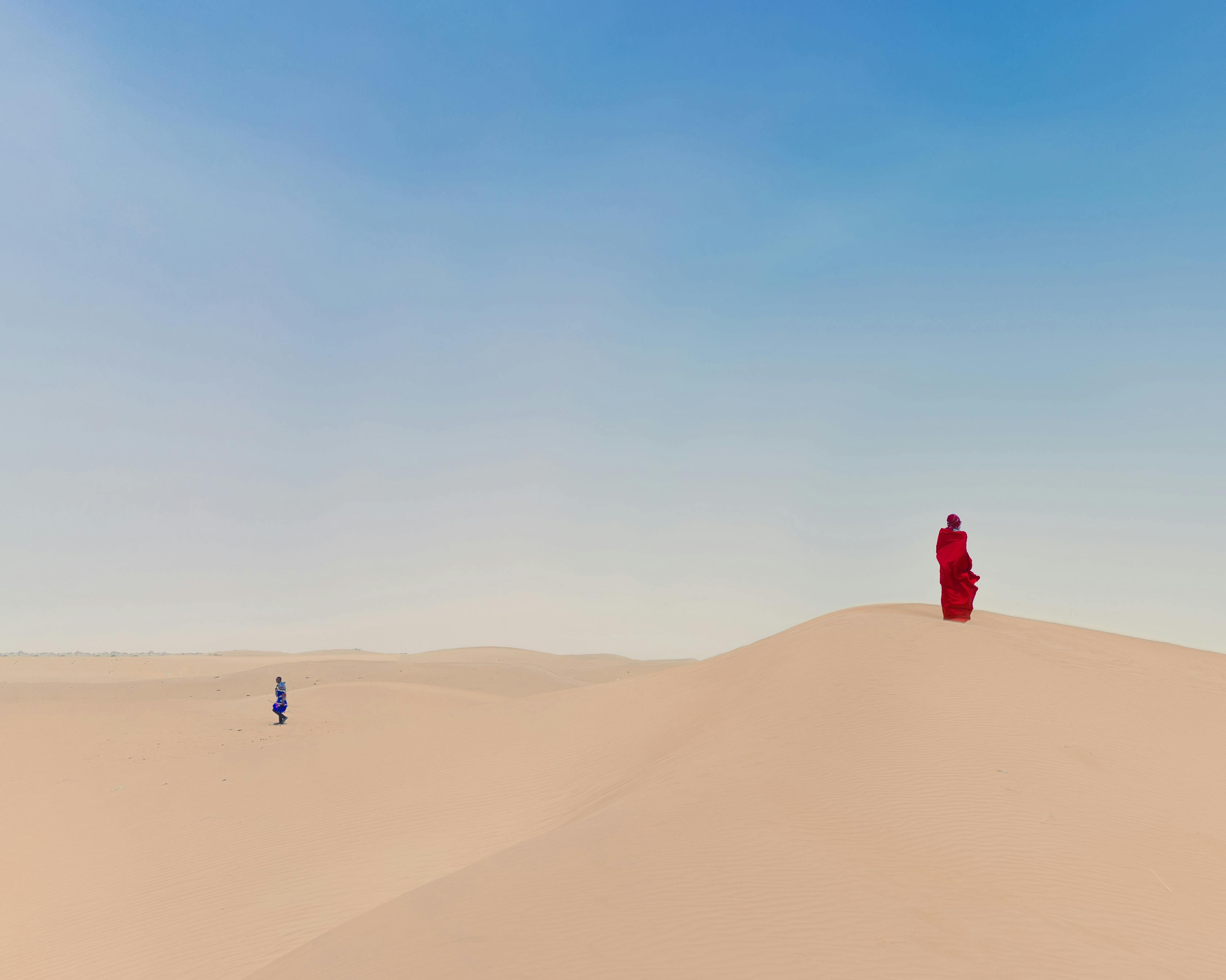 Gratuit Deux individus vêtus de vêtements aux couleurs vives se tiennent seuls dans un vaste désert, sous un ciel d'un bleu limpide. Photos
