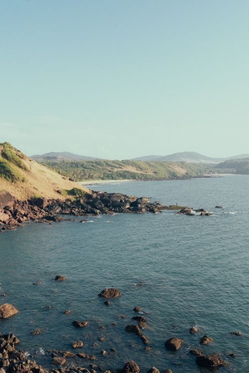 Free Beautiful coastal view with rocky shoreline and lush hills under clear skies. Stock Photo