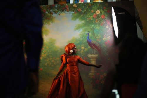 Free A model in an orange dress poses gracefully in front of a vibrant peacock backdrop in a studio setting. Stock Photo