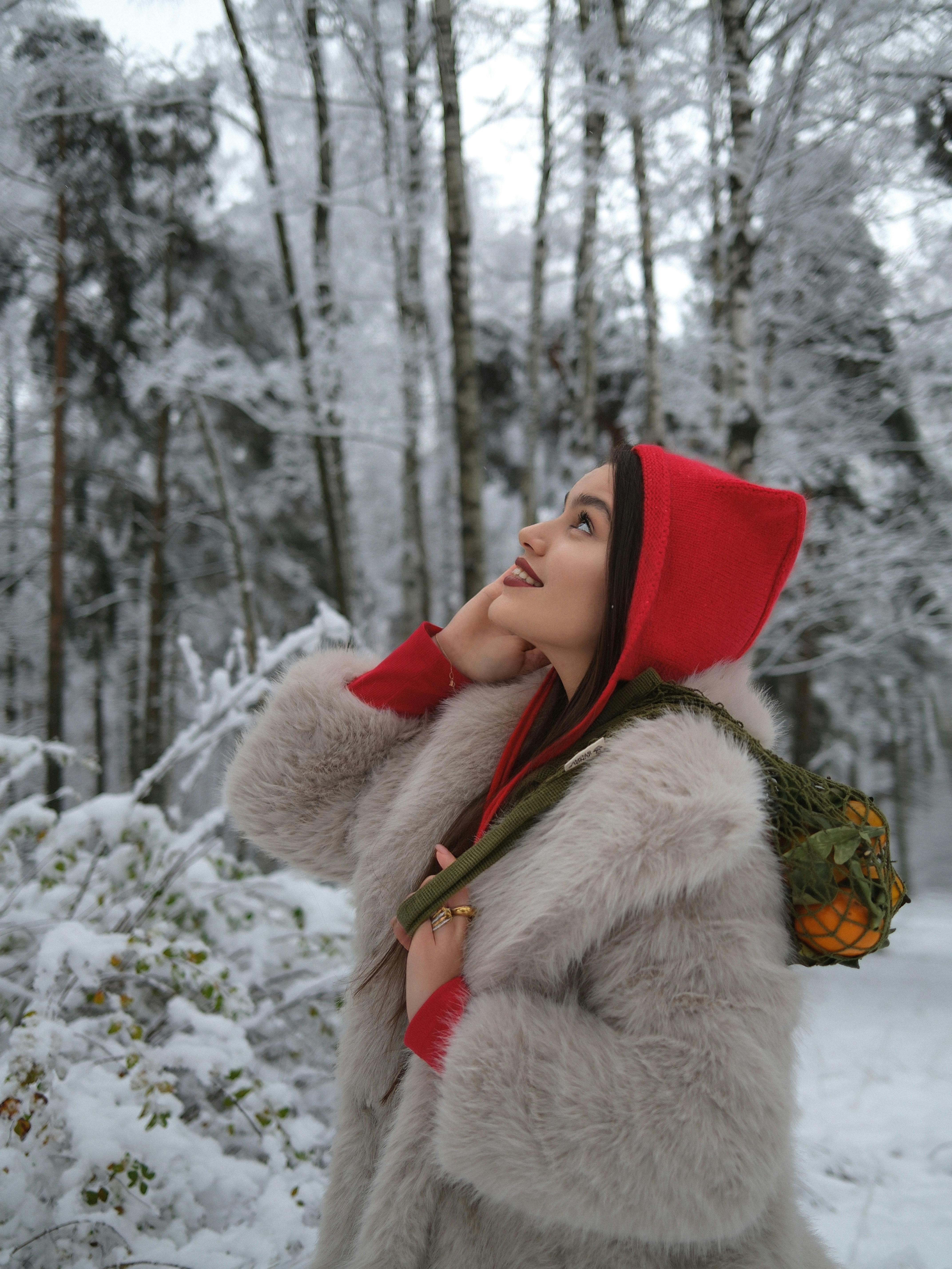 Woman wearing a red hood and fur coat in a serene snowy forest setting.