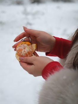 Close-up of a woman peeling a tangerine outdoors in winter snow, wearing cozy red and white.