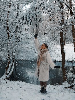 Woman in winter coat enjoying snowfall by a river in a snowy forest.
