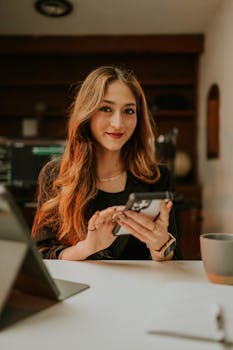 Young woman smiling while using a smartphone at a modern office desk, creating a welcoming atmosphere.