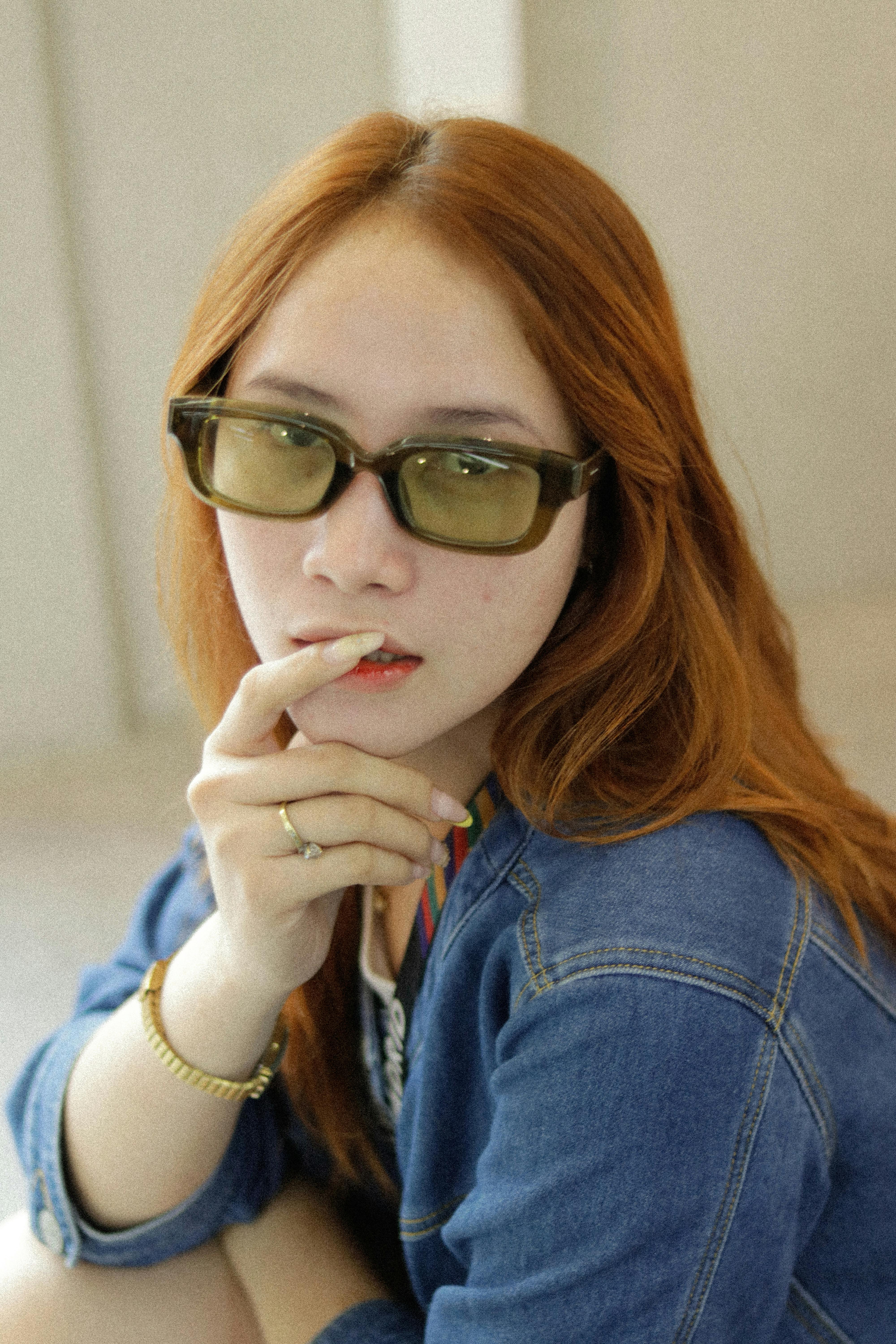 Kostnadsfria Snygg kvinna med rött hår i solglasögon och jeansjacka, poserar inomhus. Stock foto