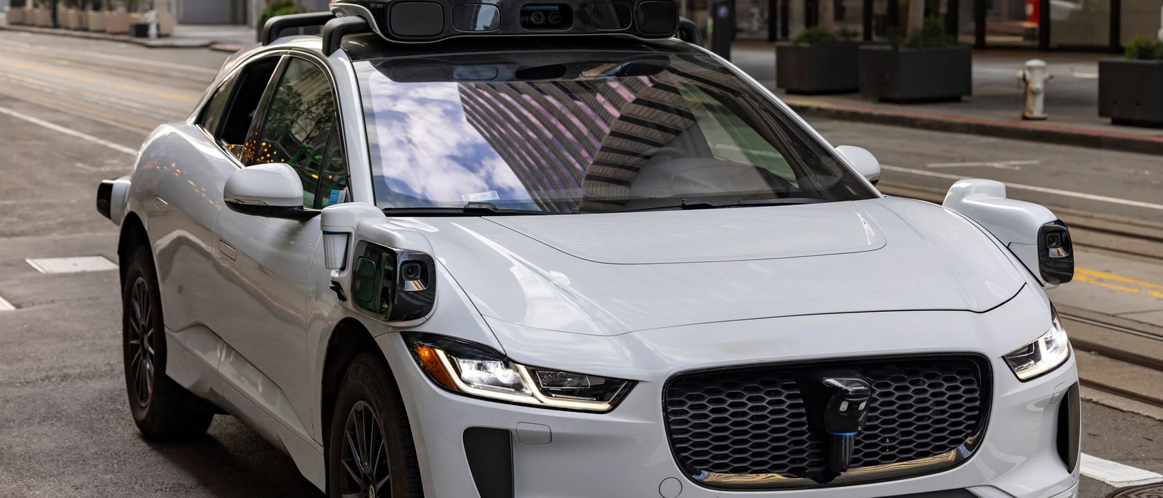 A white autonomous vehicle navigating a city street, reflecting urban architecture in daylight.