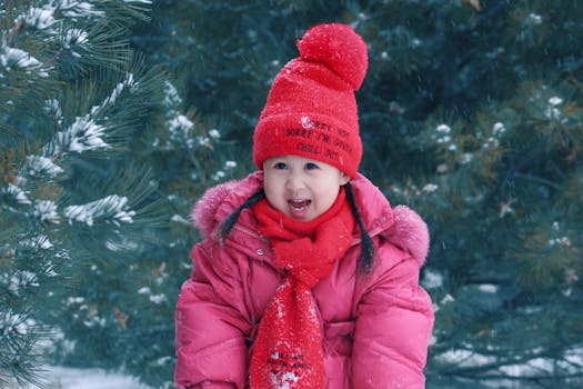 A young girl in winter attire smiles in a snowy outdoor setting, surrounded by pine trees.