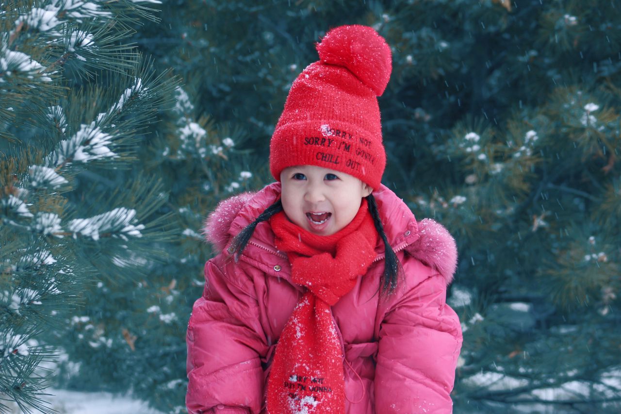 Young girl in a pink puffer coat, red pom-pom hat and red scarf in a snowy pine forest