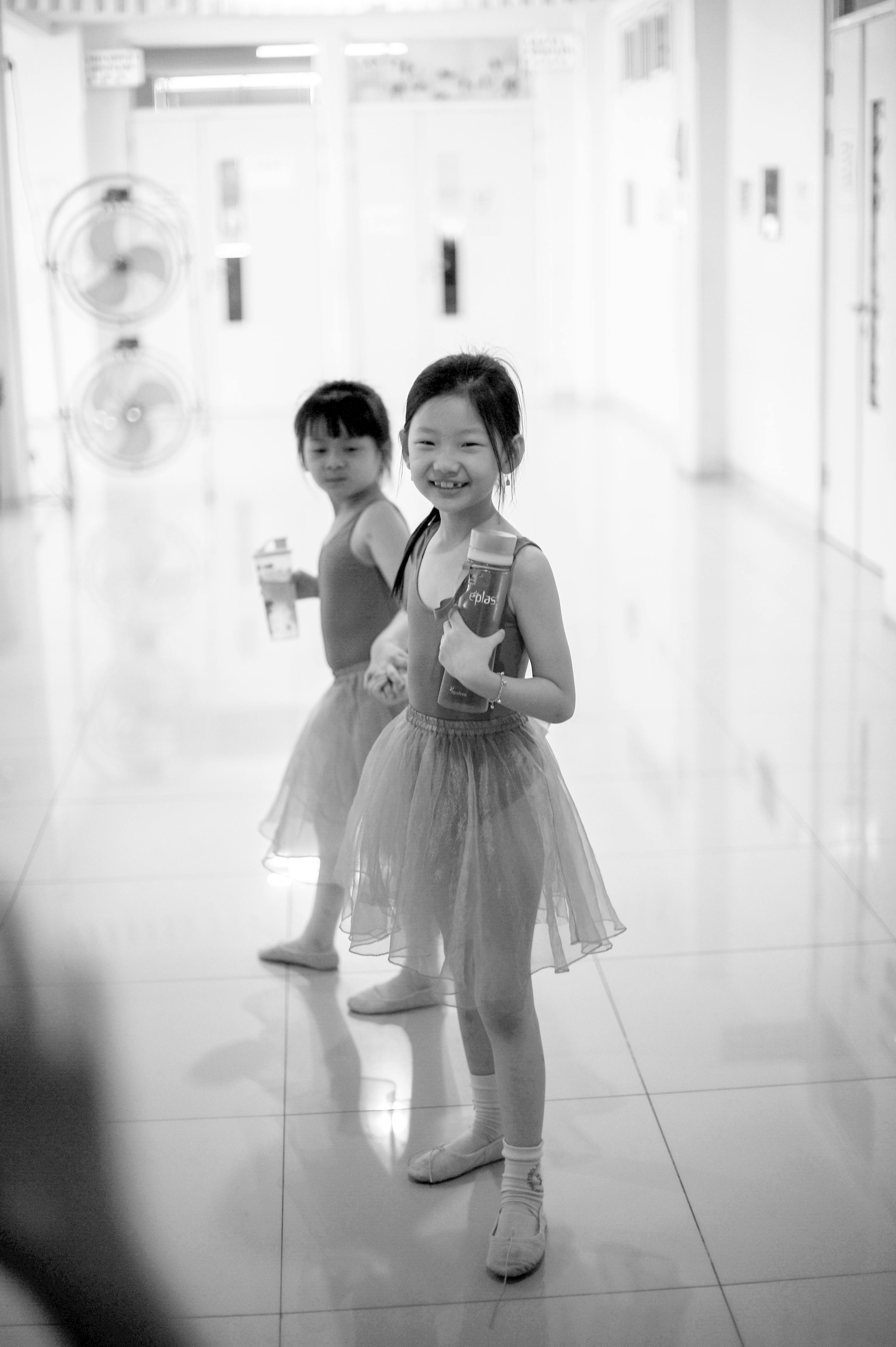 Two young girls in ballet attire smiling and holding drink cartons in a dance studio.