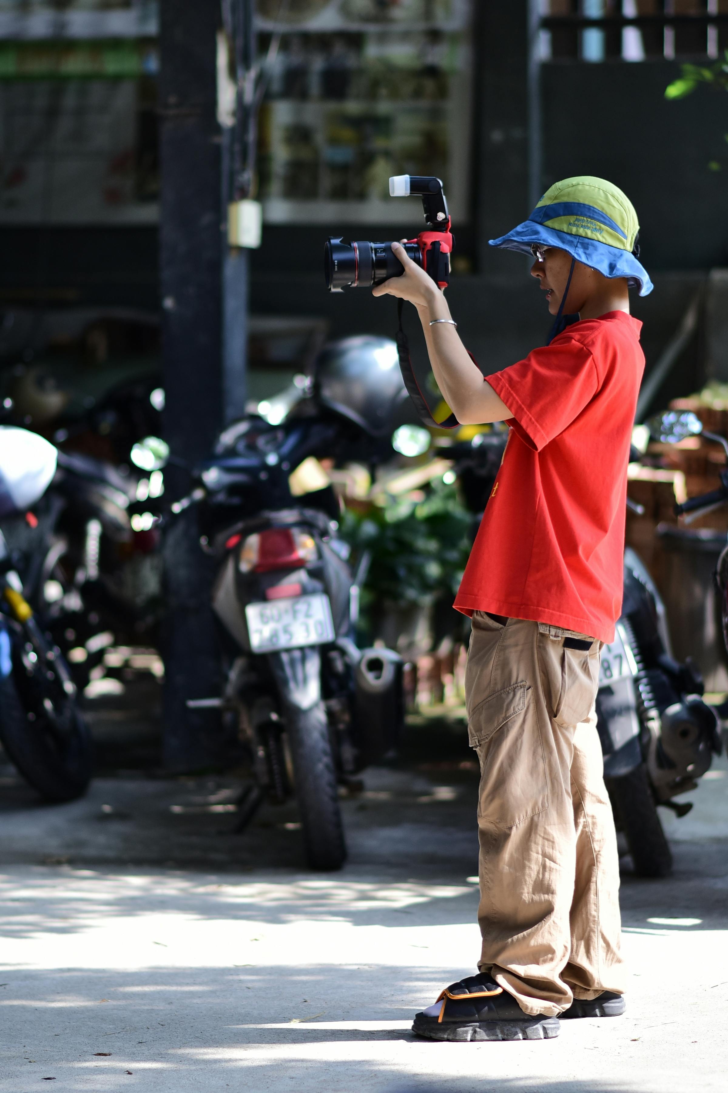 A young photographer in a red shirt and blue hat takes pictures outdoors with a DSLR camera.