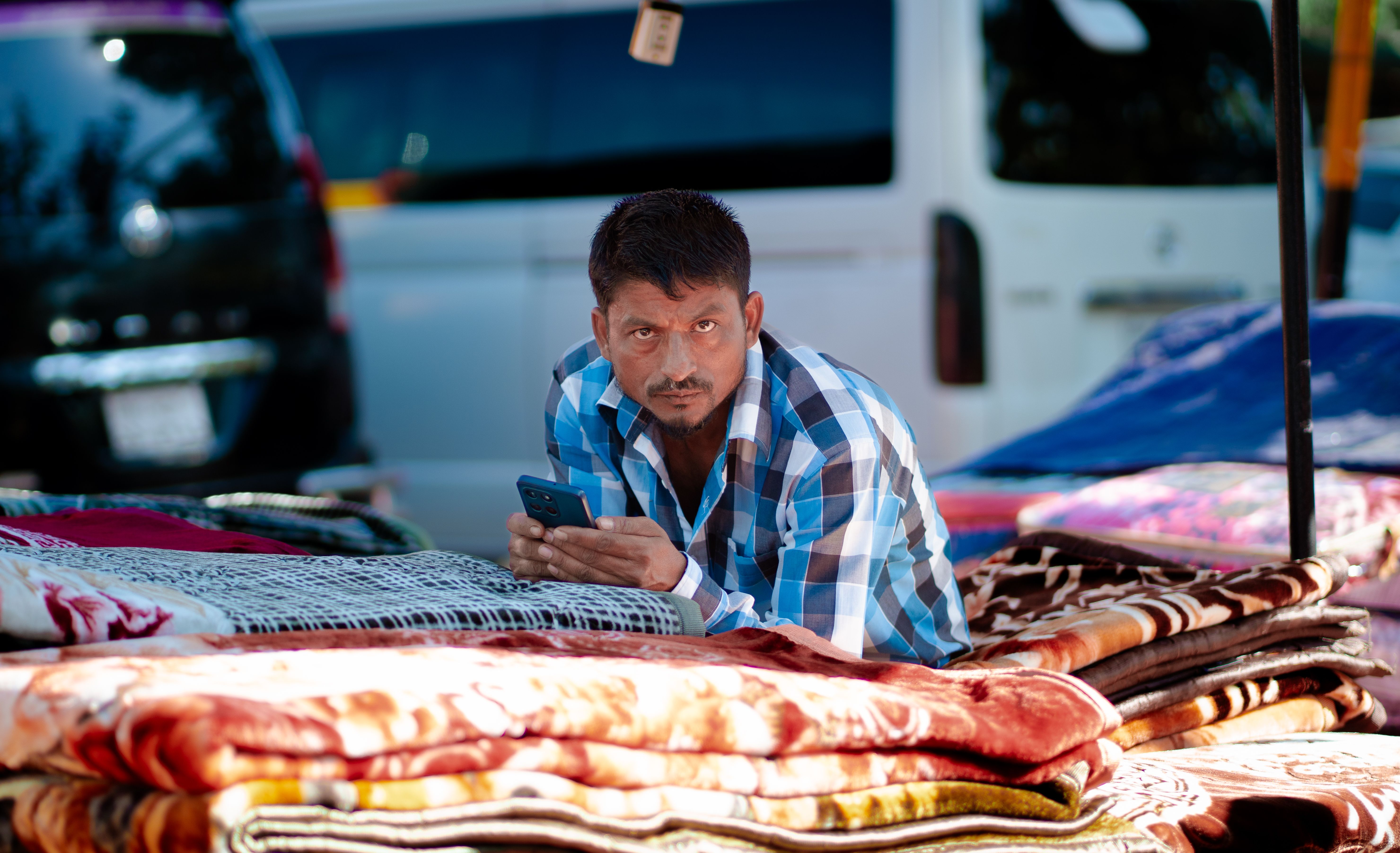 Dark-skinned Nigerian male trader in a plain white kaftan using a smartphone to process a payment at a colourful Oyo State open-air food market stall, fresh tomatoes, peppers, and bags of garri on the table in front of him, other market stalls visible in the background, bright natural daylight, photorealistic