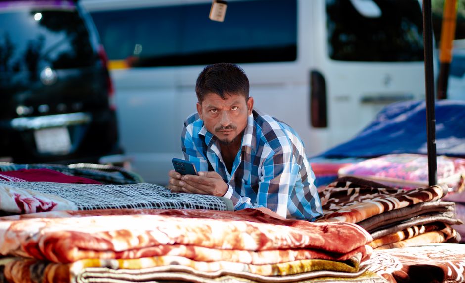 Dark-skinned Nigerian male trader in a plain white kaftan using a smartphone to process a payment at a colourful Oyo State open-air food market stall, fresh tomatoes, peppers, and bags of garri on the table in front of him, other market stalls visible in the background, bright natural daylight, photorealistic
