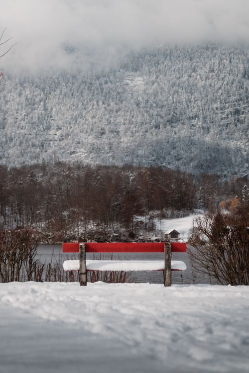 Free Peaceful snowy scene featuring a red bench overlooking a tranquil lake and misty mountains. Stock Photo