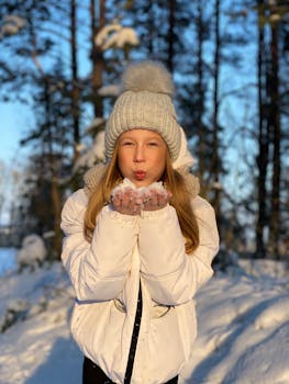 Young girl in white jacket blowing snowflakes in a sunny winter forest.