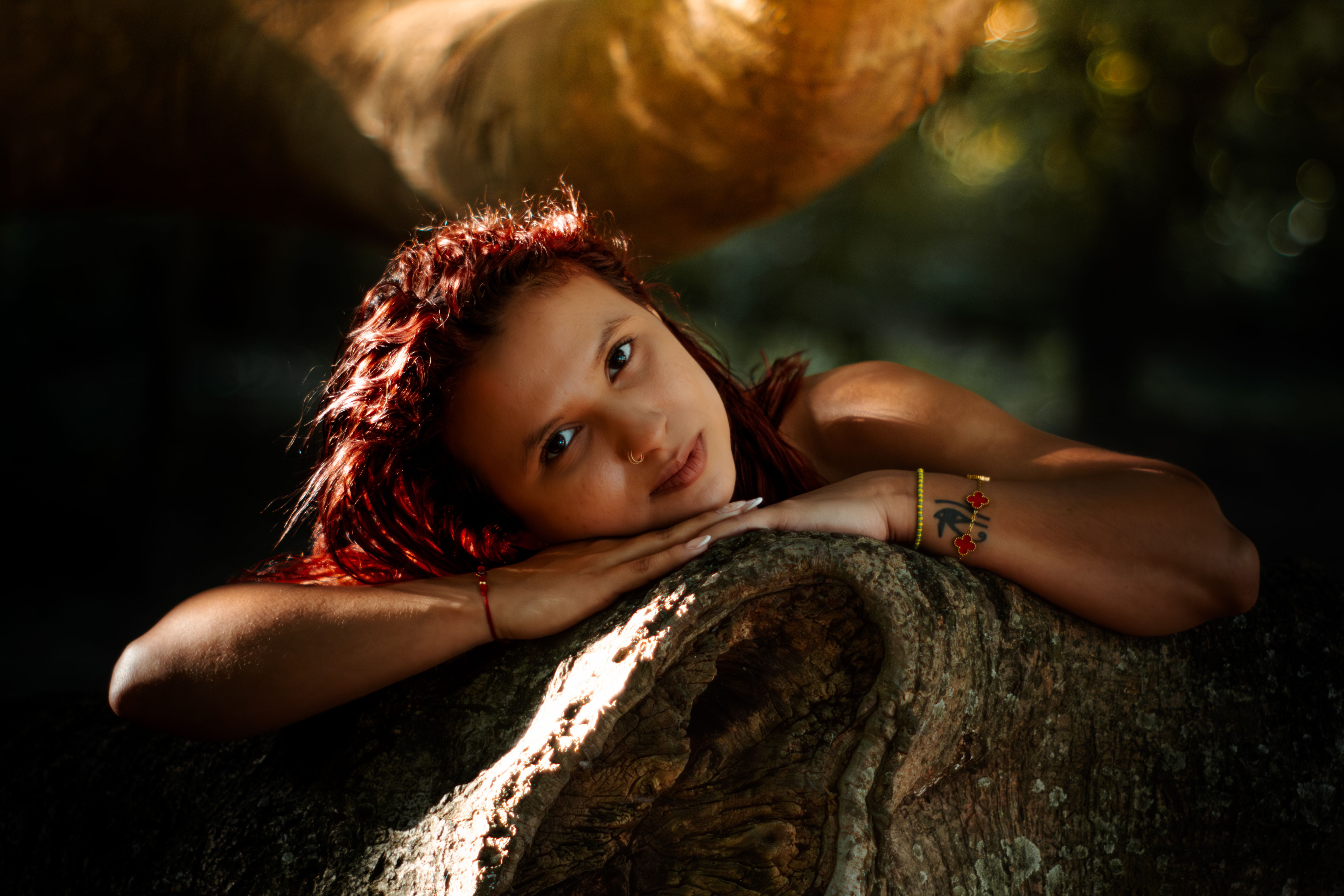 A woman with red hair rests peacefully on a tree trunk in a sunlit forest setting.