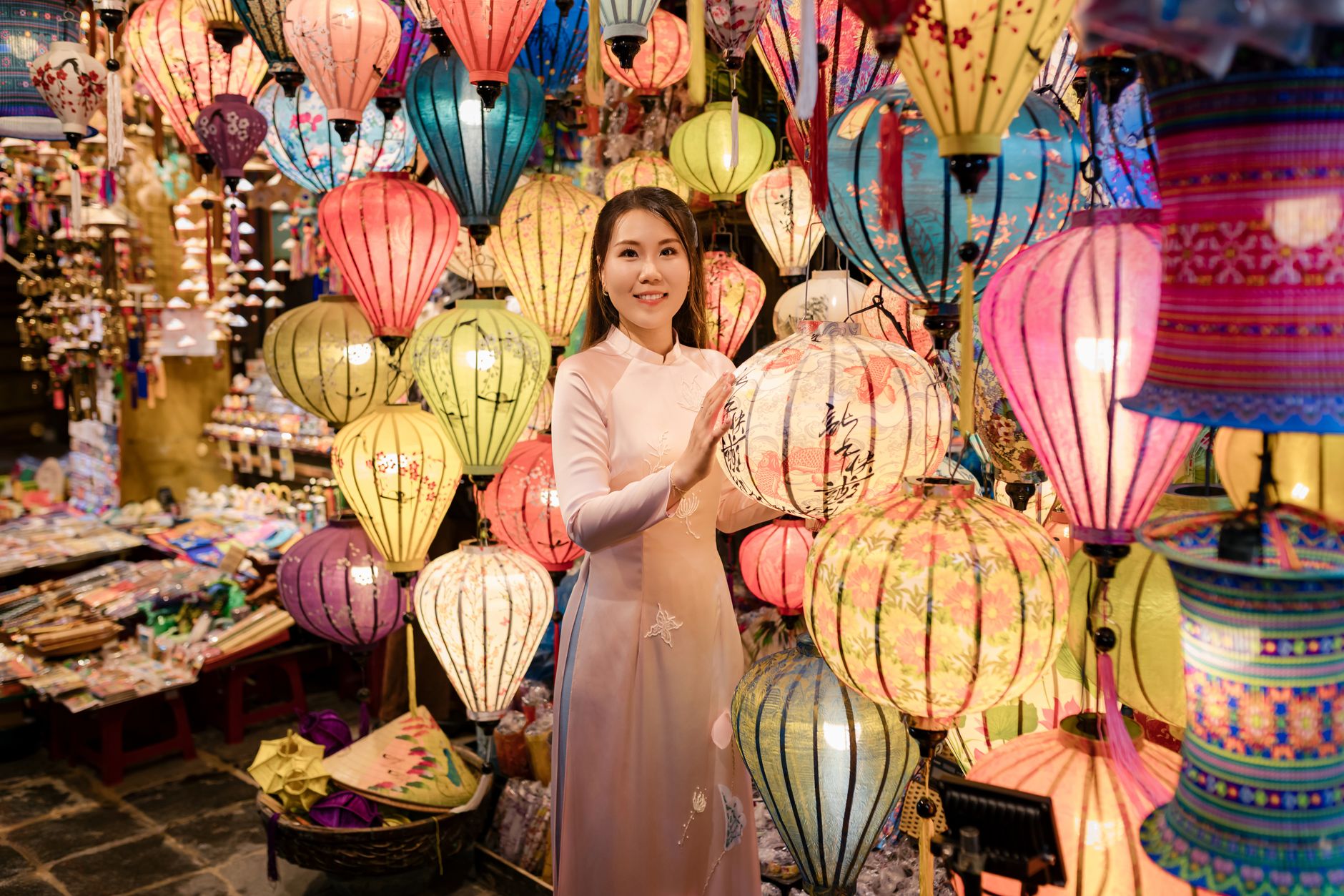 A woman in traditional Ao Dai surrounded by vibrant lanterns at a market in Hội An, Vietnam at night.