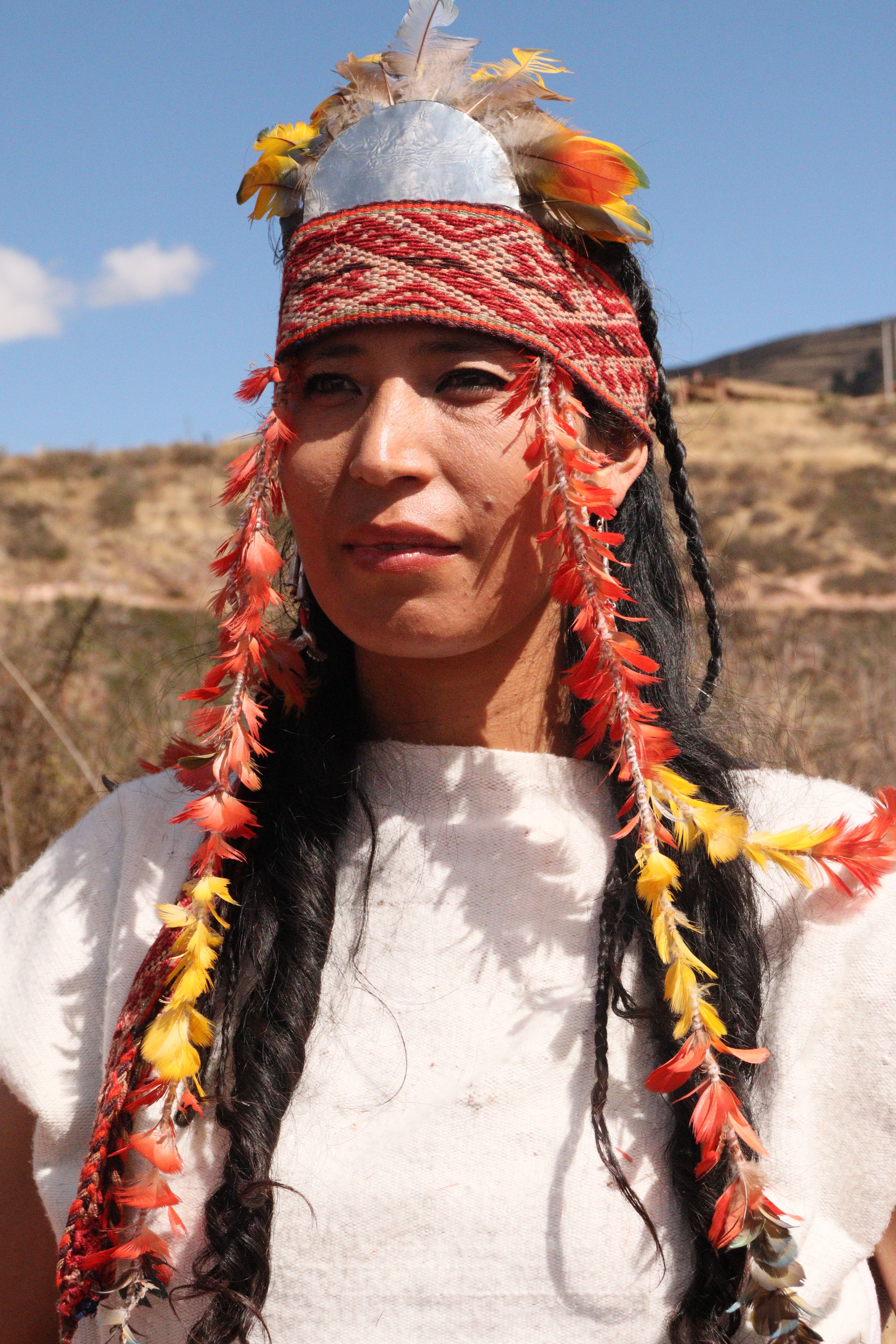 A person wearing traditional attire with feather adornments, outdoors under a sunny sky.