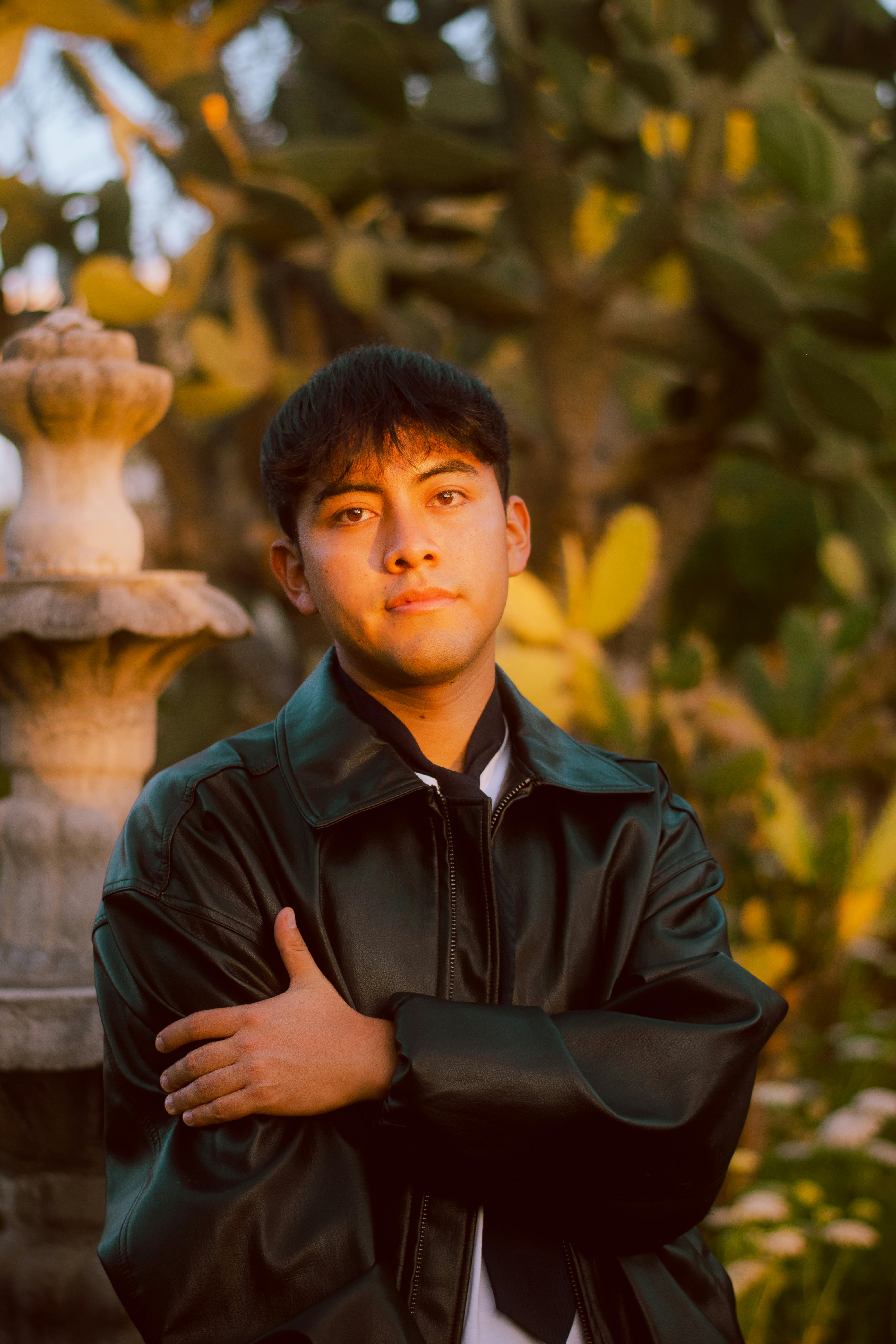 Portrait of a young man in a black leather jacket surrounded by lush greenery and sunlight.