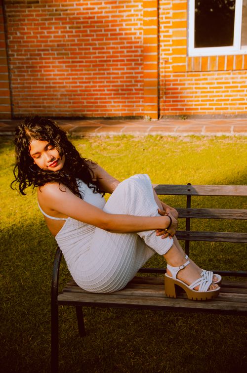 Free Woman in a white dress sitting on a bench with brick wall background. Sunlit and serene. Stock Photo