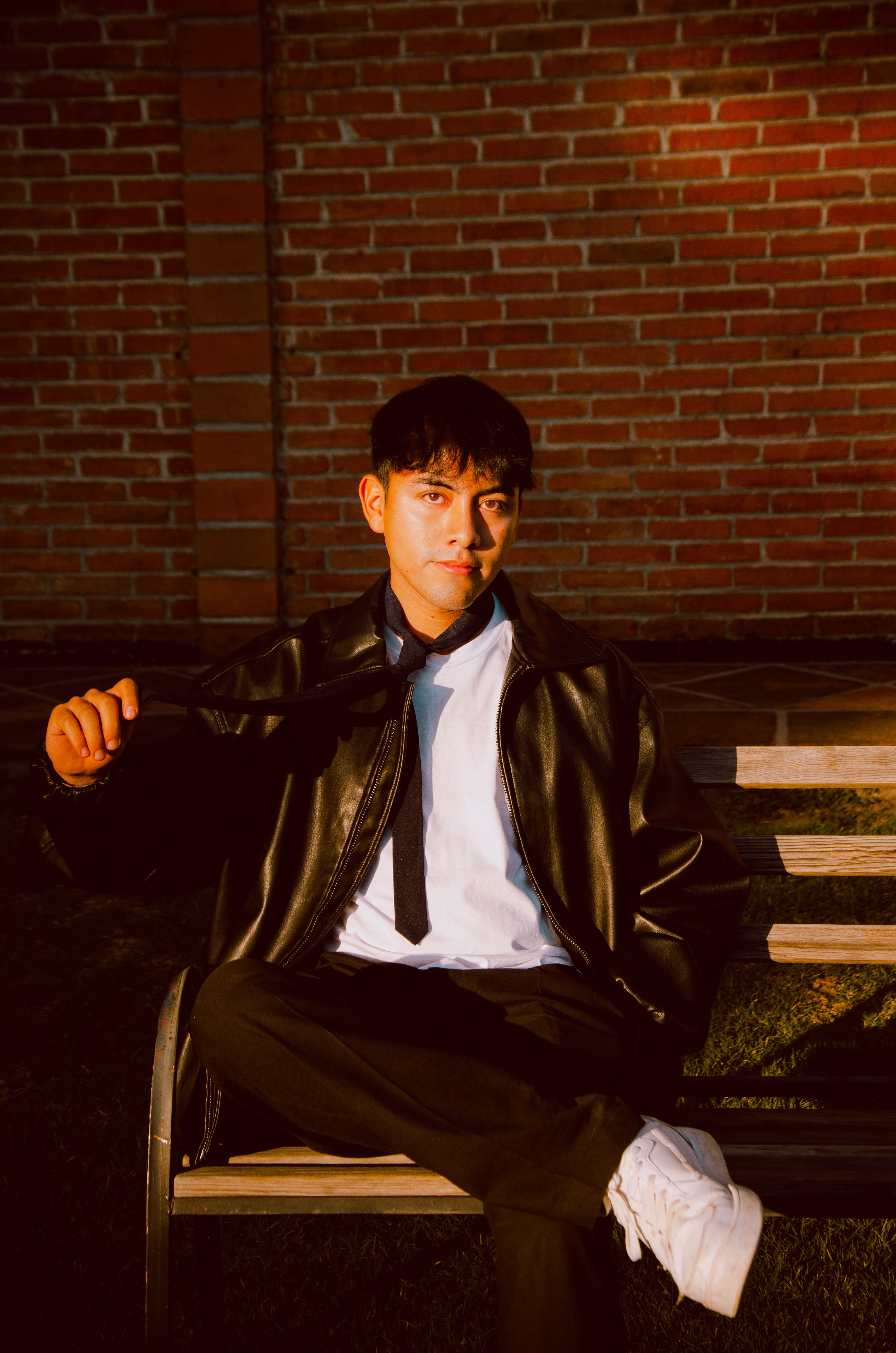 A stylish young man in a leather jacket sits on a bench against a brick wall at sunset.