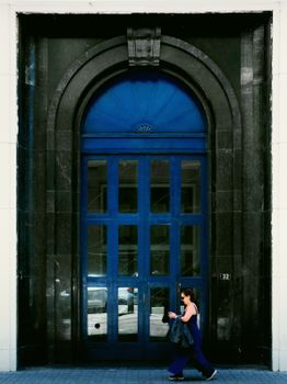 Stylish woman walks past an ornate blue door on a sunny day.