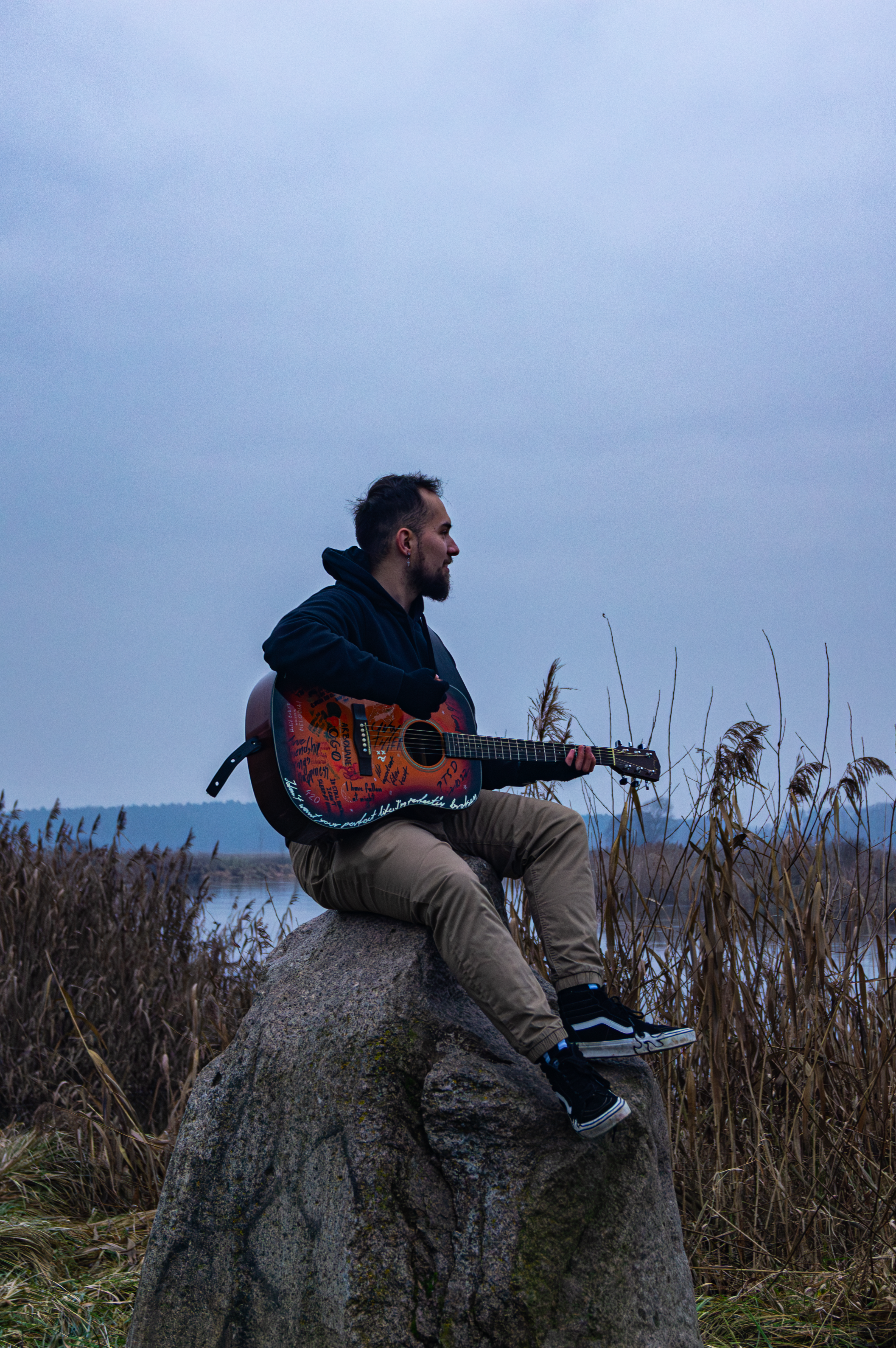 A man playing acoustic guitar on a rock beside a lake, serene autumn setting.