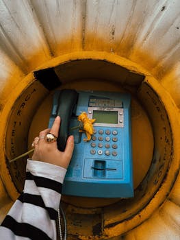 A hand holding a retro blue payphone receiver with a yellow flower, set in a vibrant yellow booth.