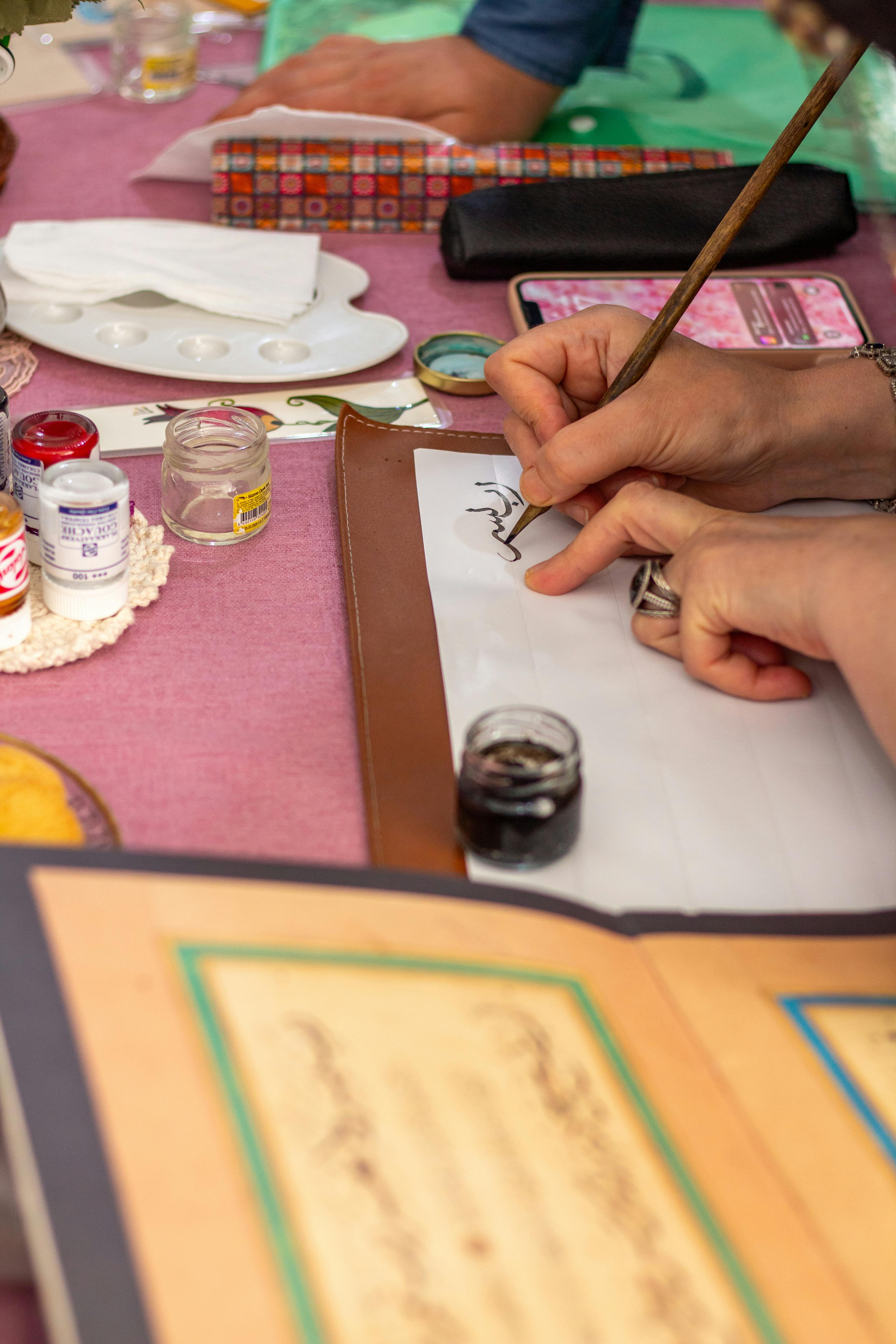 Hands writing calligraphy with a quill and ink on a table with art supplies.