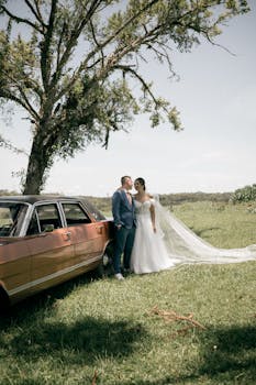 Bride and groom share a moment outdoors beside a vintage car under a tree.