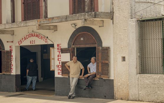 Two men talking outside a vintage parking garage in an urban setting.