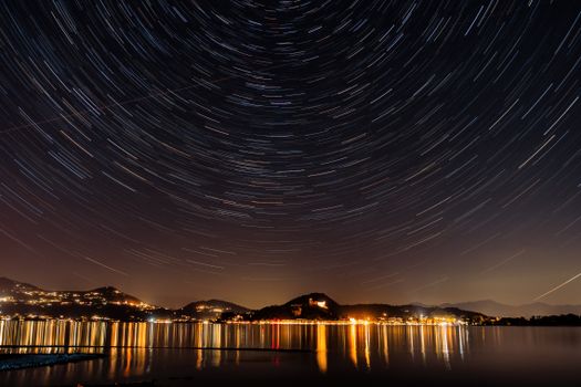 Star trails over a calm lake reflecting city lights, creating a serene and dramatic night scene.