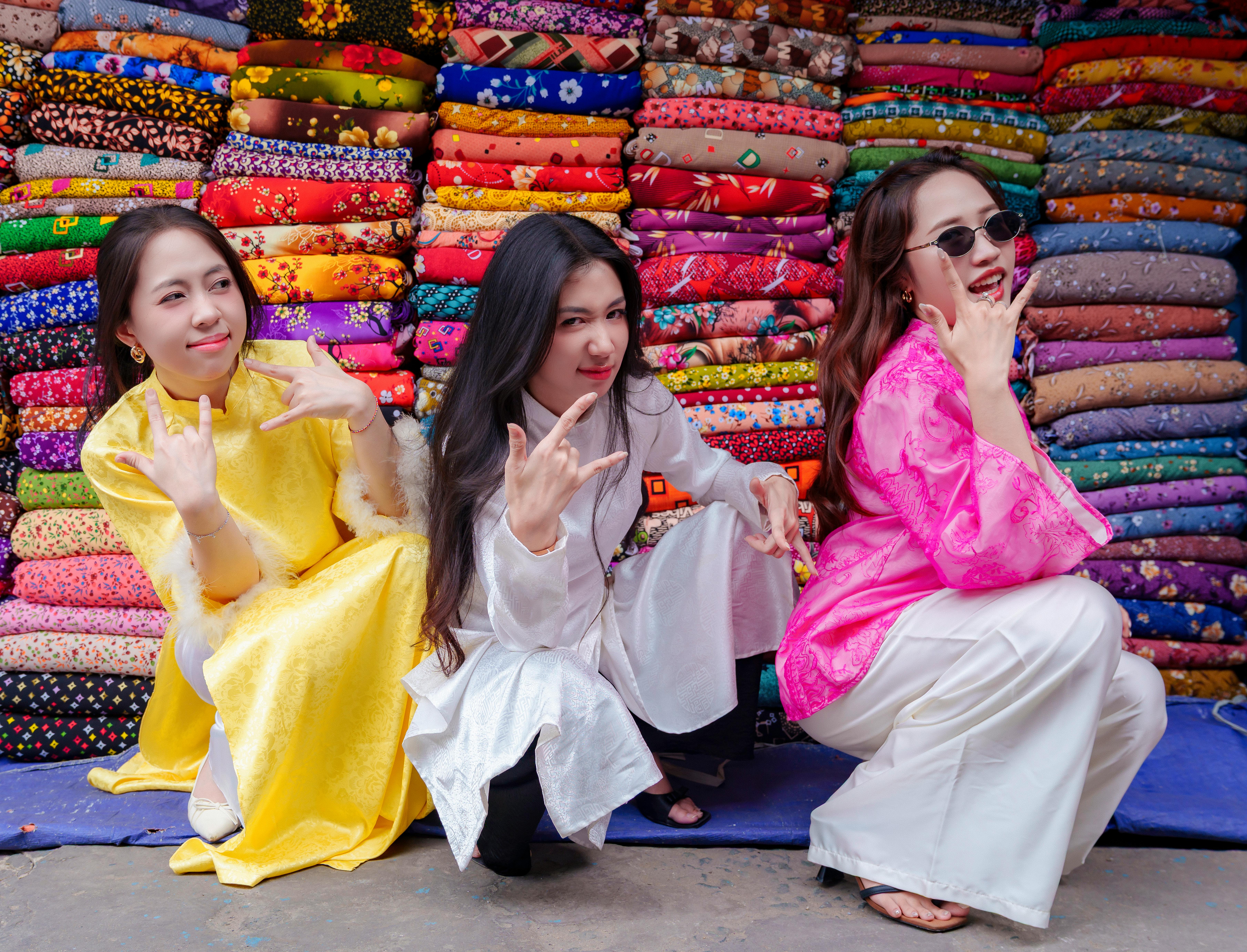 Three women in colorful dresses pose playfully in front of fabric bundles in Ho Chi Minh City.
