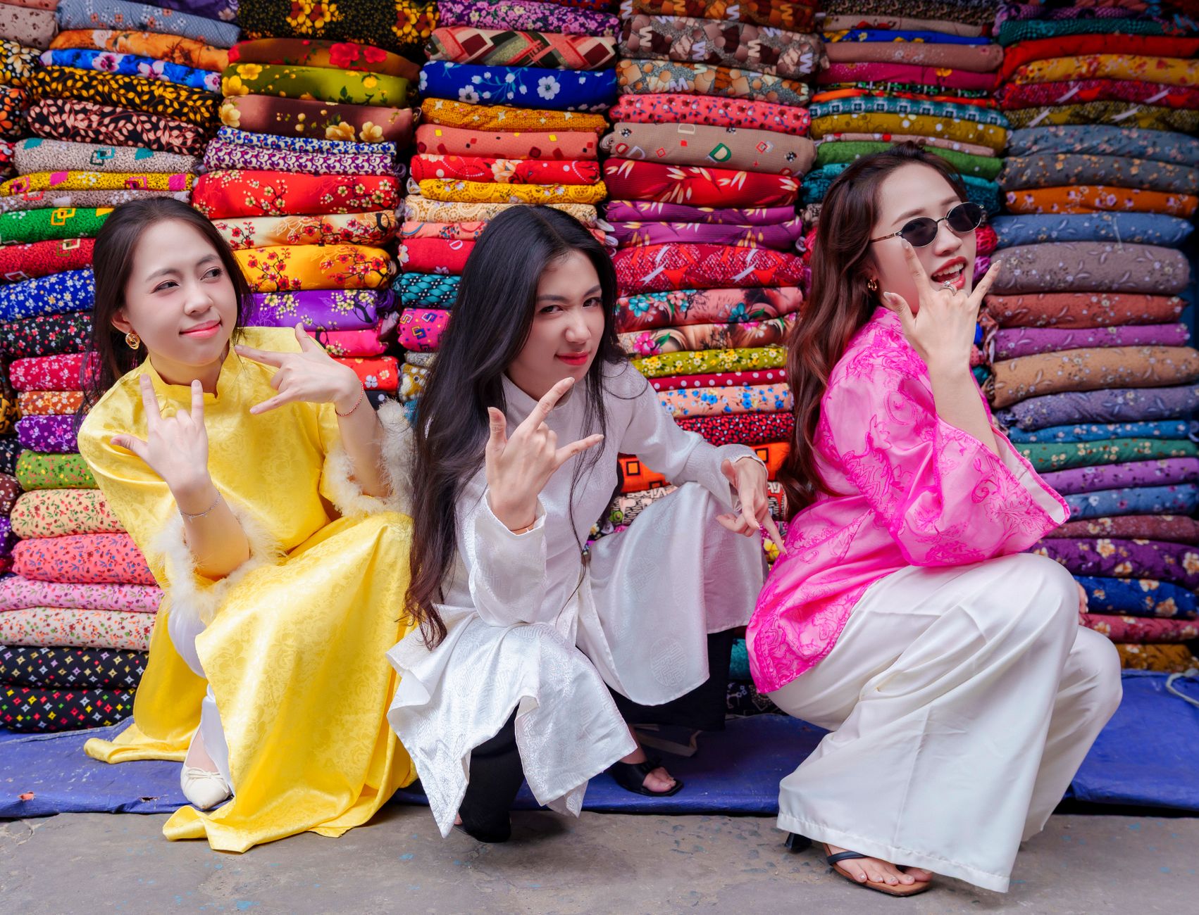 Three women in colorful dresses pose playfully in front of fabric bundles in Ho Chi Minh City.