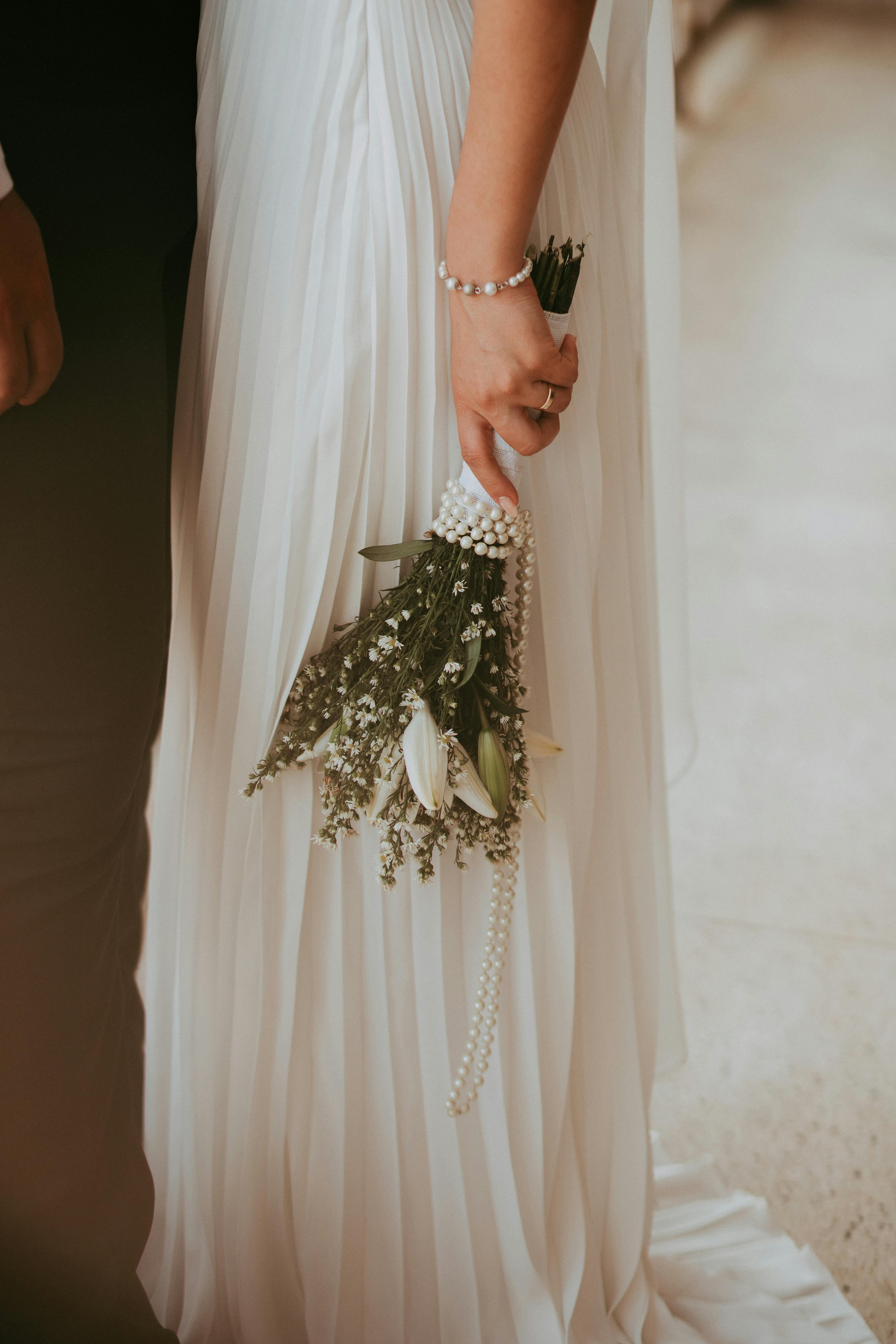 Close-up of a bride holding a delicate bouquet with white roses and greenery, wrapped in pearls.