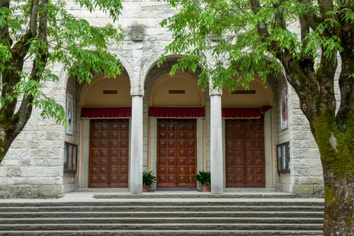 Free An elegant stone building facade with large wooden doors and archways framed by lush green trees. Stock Photo