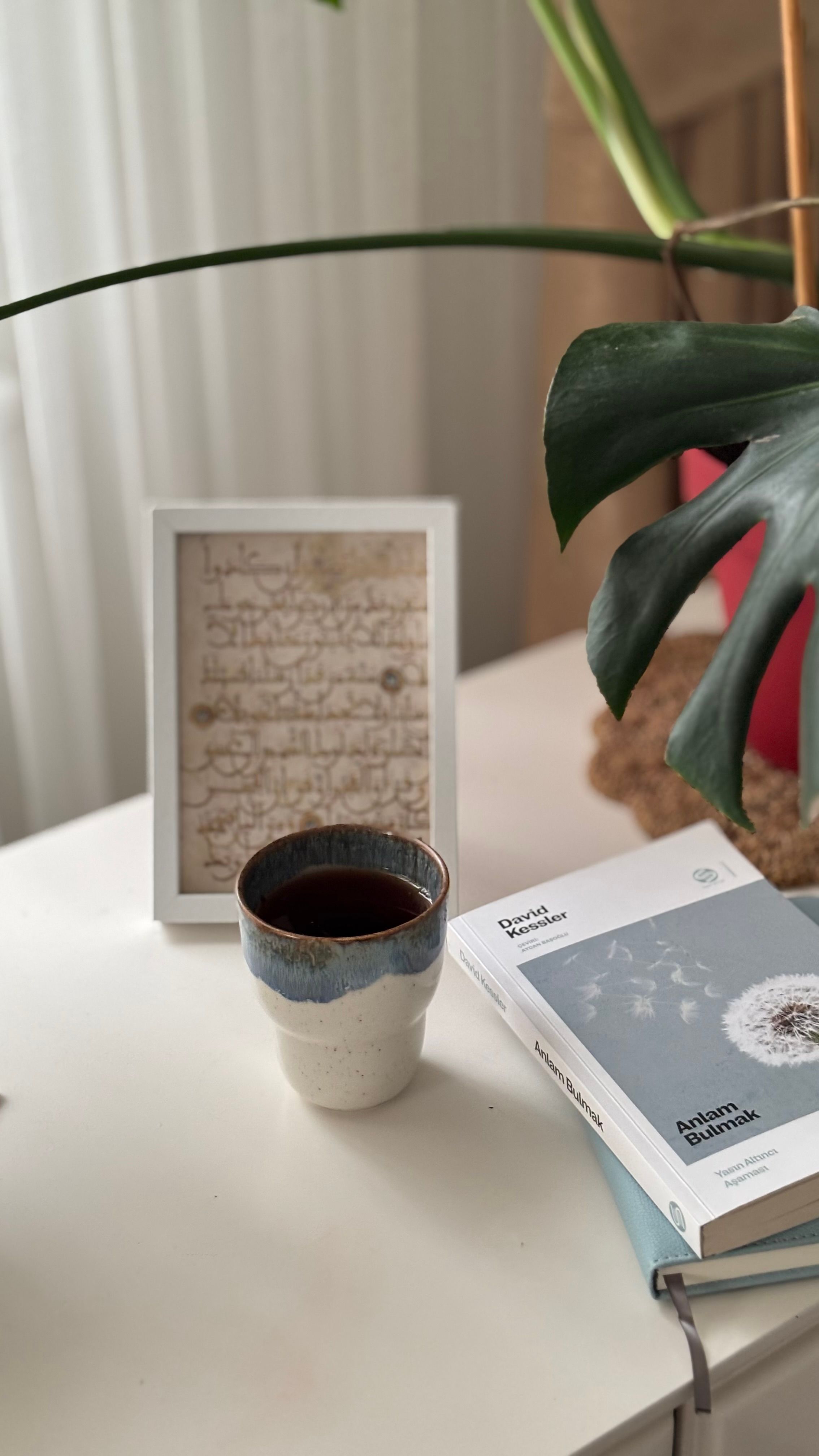 A serene reading corner with a ceramic mug, plant, and book on a white table.