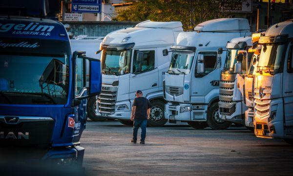 Truck Hire (with driver) โ A man walks among parked trucks at sunset in an industrial area, creating a serene yet busy scene.