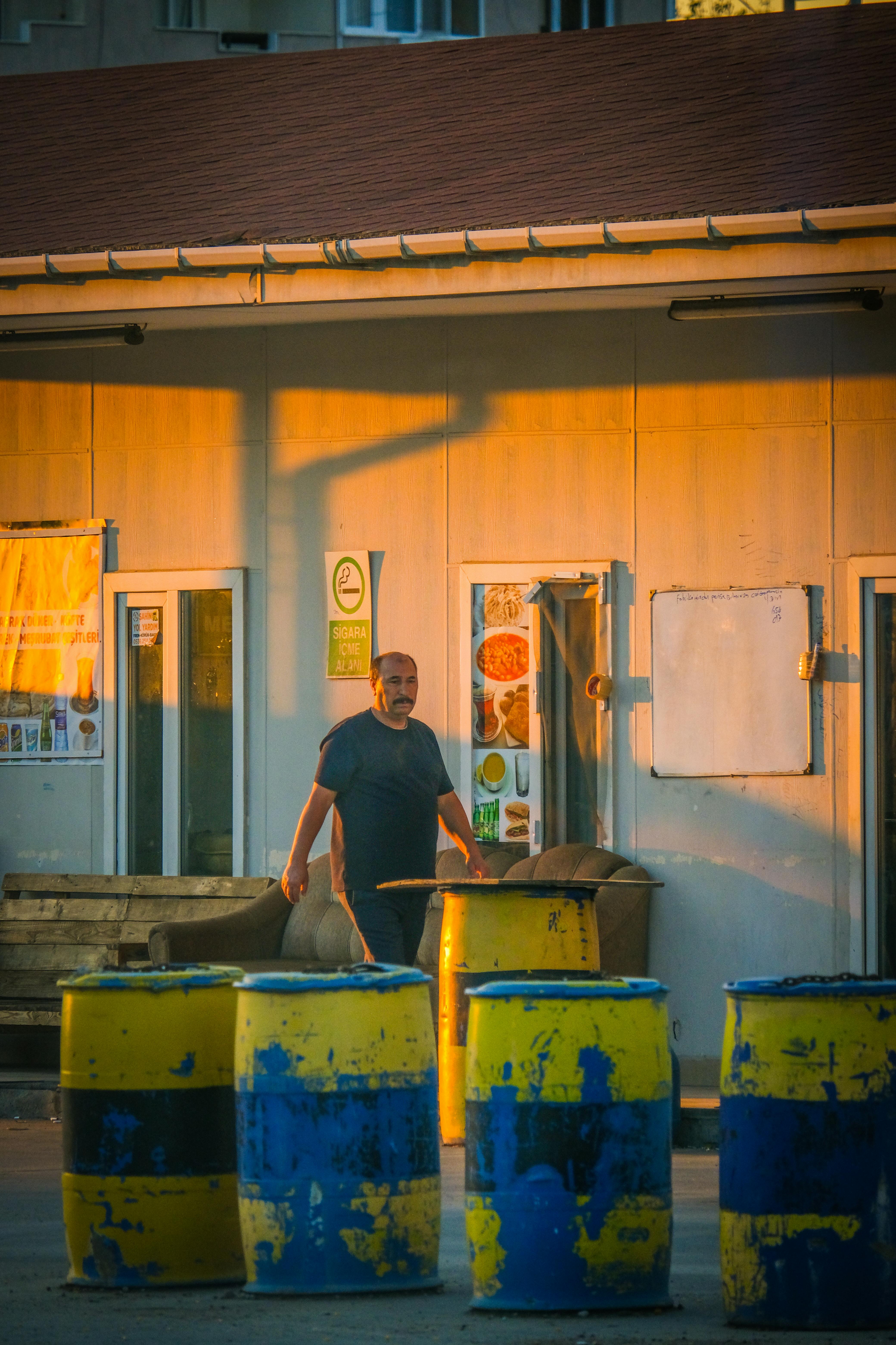 Безкоштовне Чоловік стоїть біля синіх та жовтих бочок біля будівлі на заході сонця. стокове фото