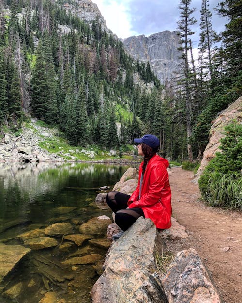 Free Side view of female tourist in red coat and cap sitting on rocky coast of river observing mountains in coniferous trees around Stock Photo