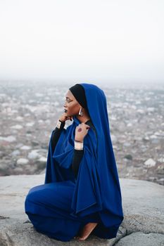 Side view of a woman in a blue hijab sitting on a rock overlooking a city, evoking a sense of peace and contemplation.