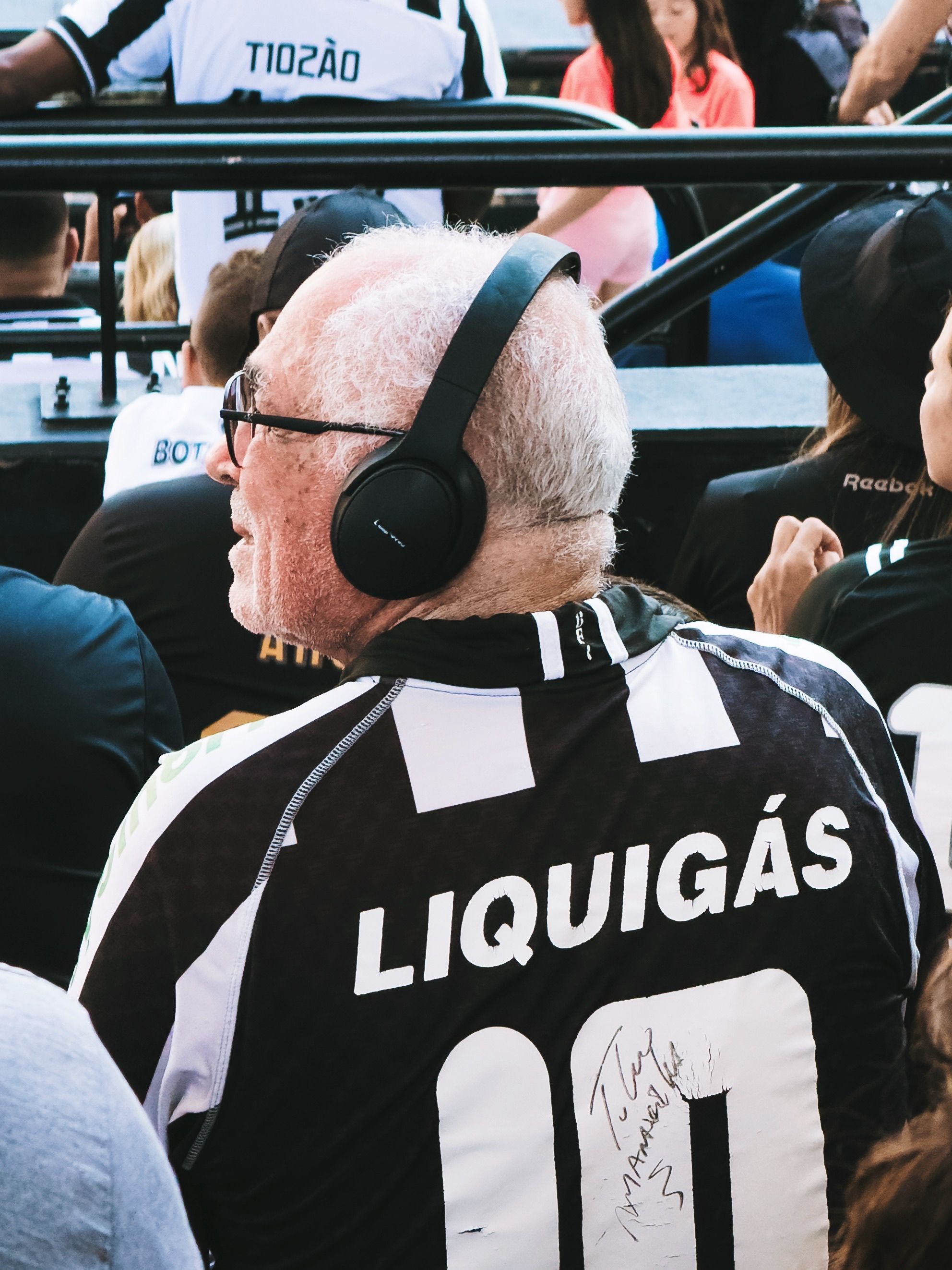 Senior man wearing jersey and headphones, enjoying a live sports event in a crowded stadium.