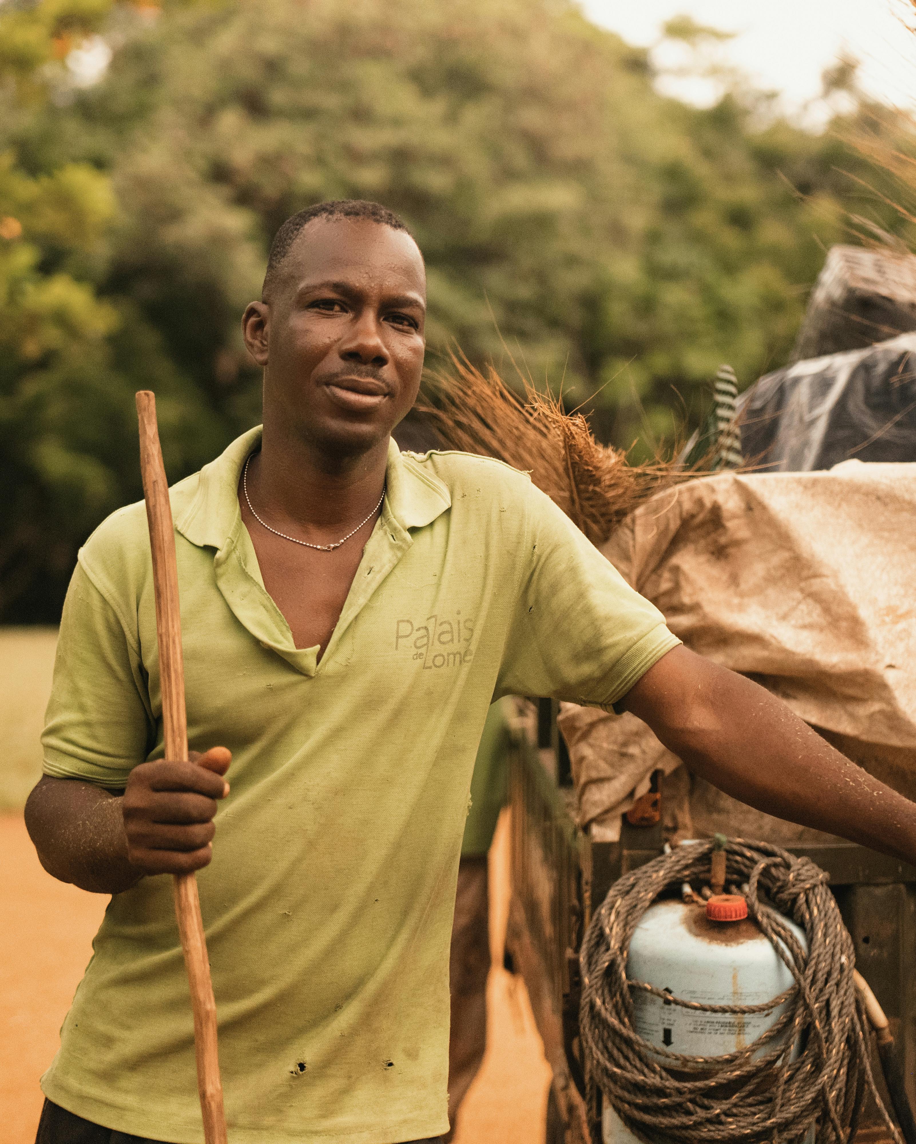 A farmer with a wooden stick stands by a cart loaded with supplies outdoors.