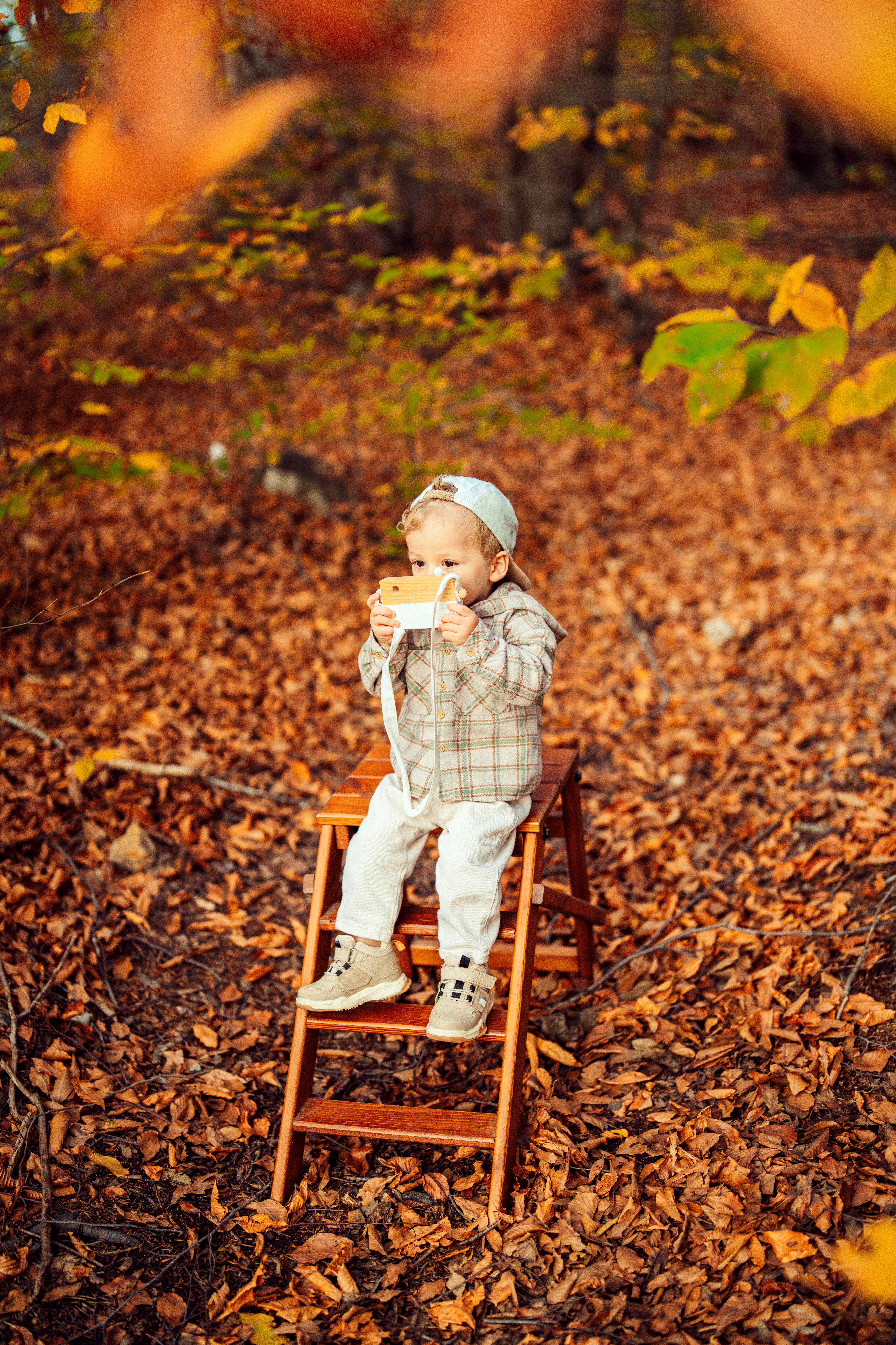 A young child sits on a wooden stool in a fall forest, capturing memories with a vintage camera.