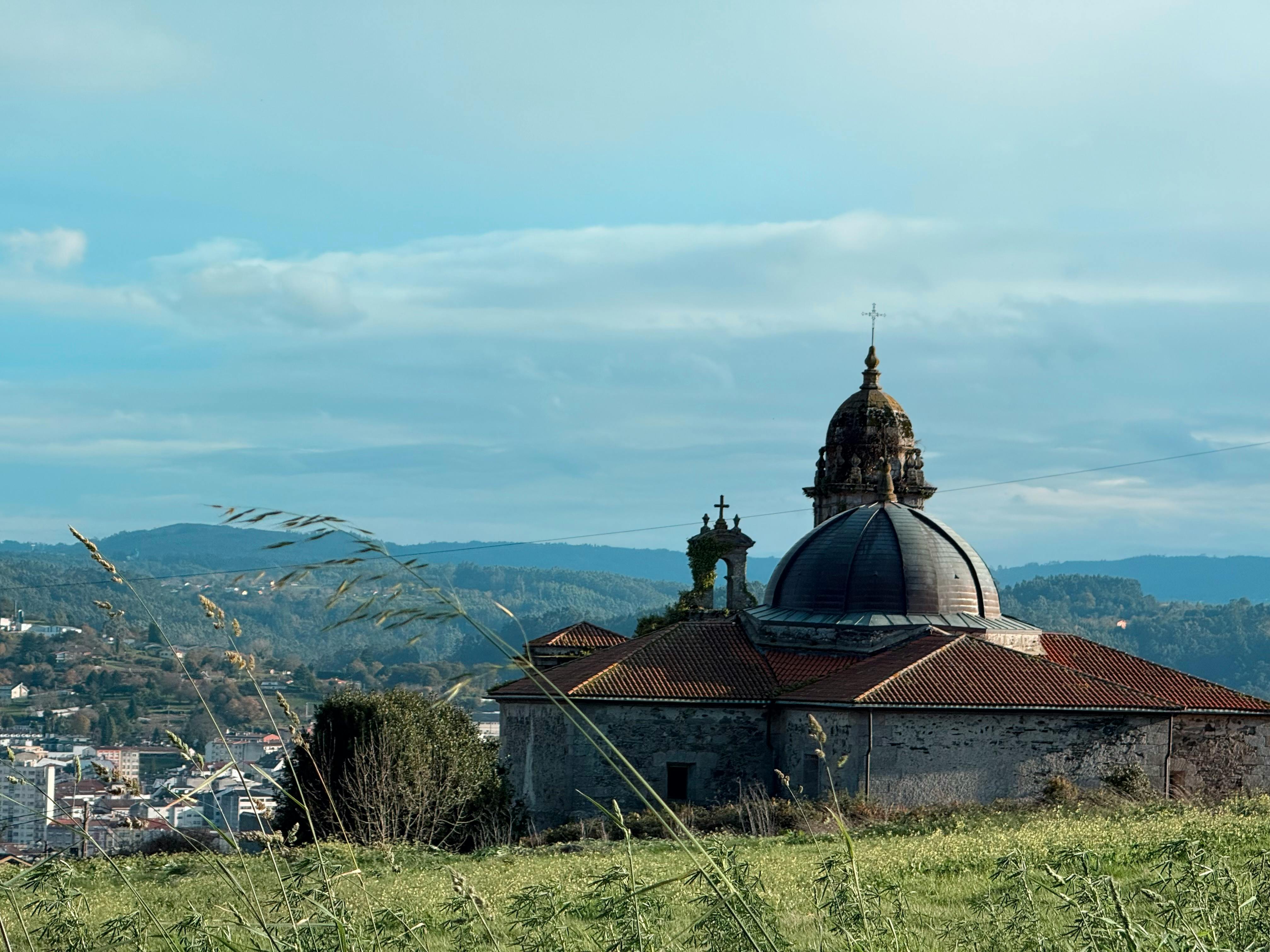 A historic church dome set against a tranquil Galician landscape under a blue sky.