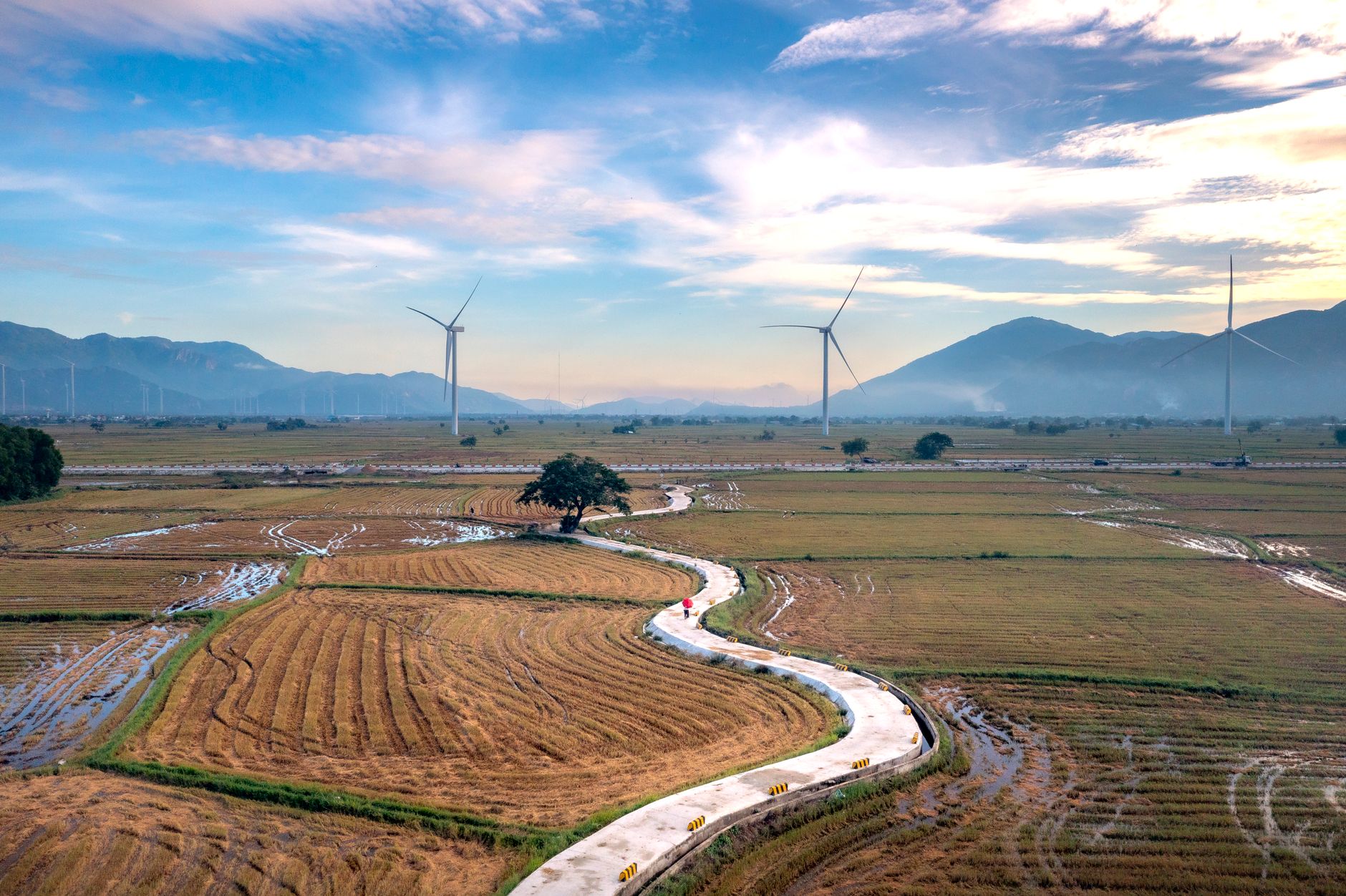 Aerial view of a winding road through fields with wind turbines and mountains in the background.
