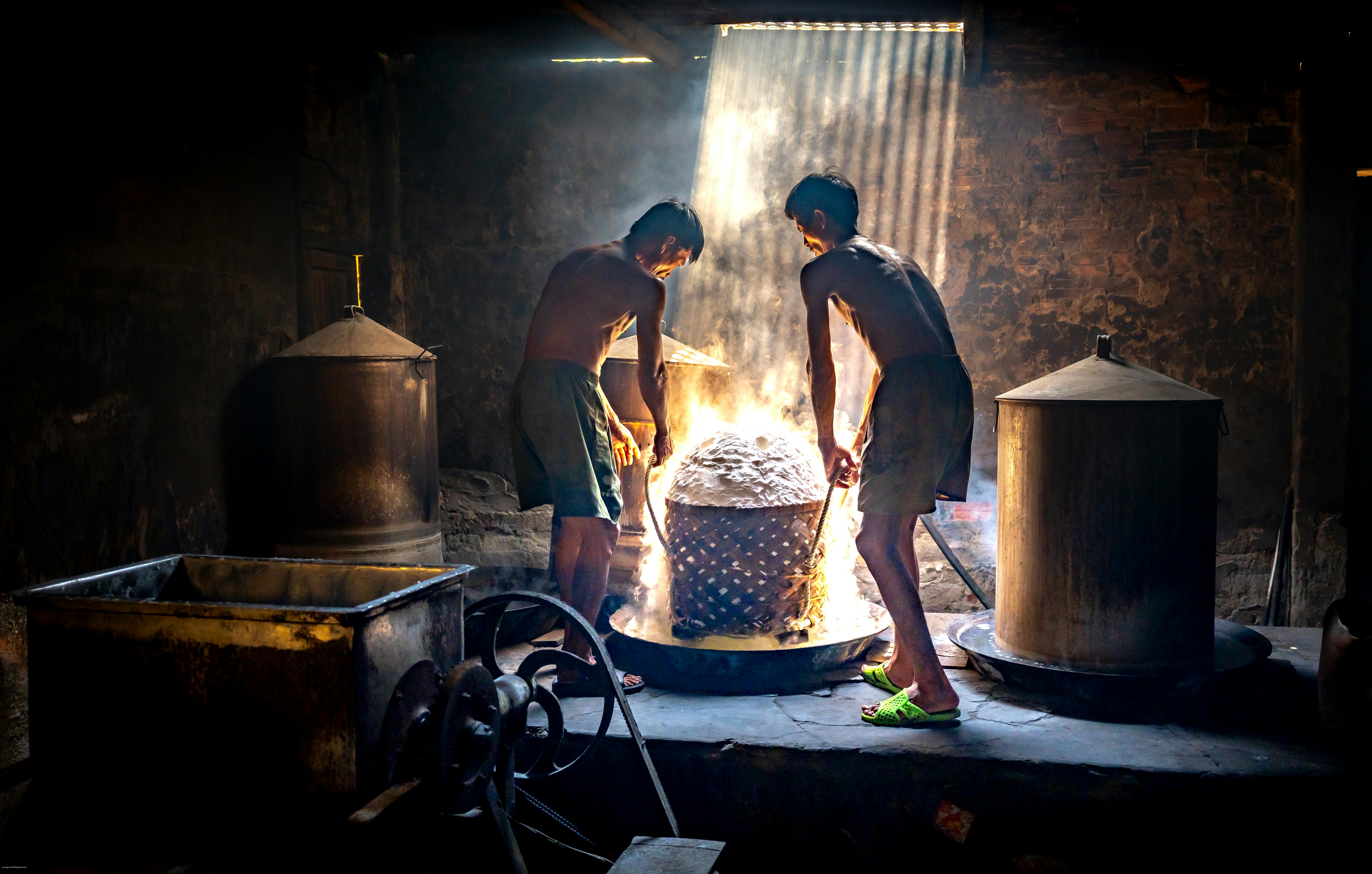 Two workers in a traditional brewery stir a vat in a dimly lit room, lit by sunlight rays.