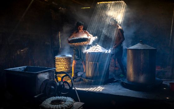 Two men create traditional food in a rustic, dimly lit workshop illuminated by sunbeams, highlighting artisanal craftsmanship.