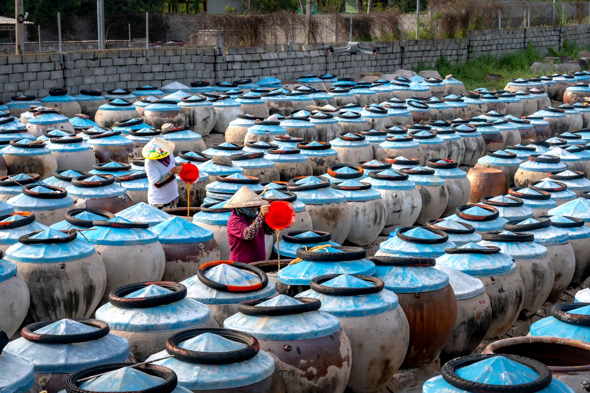 Two workers tend to large fermentation pots in an outdoor market setting, showcasing traditional methods.