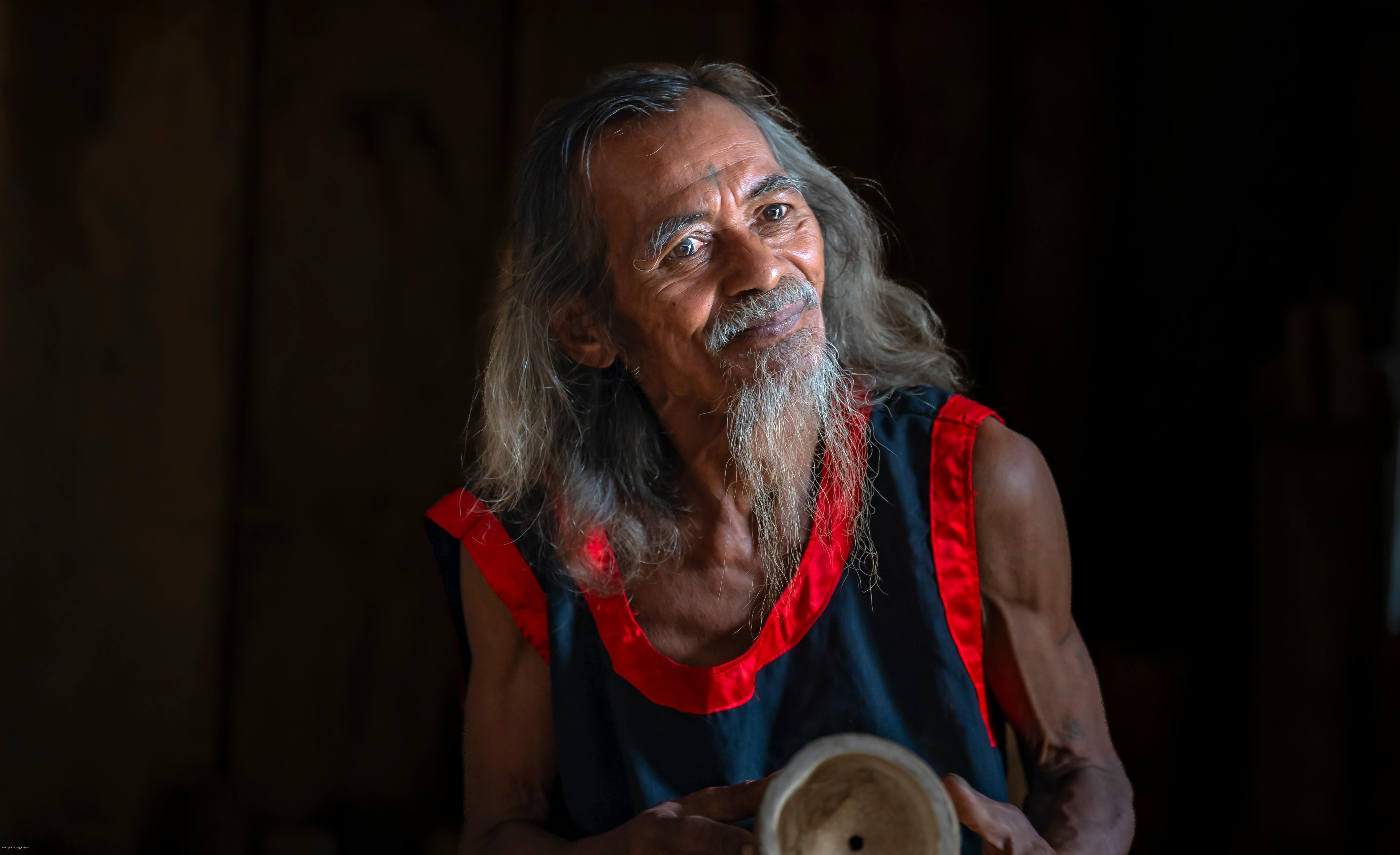 An elderly man with long hair and a beard stands indoors holding an object, with warm lighting.