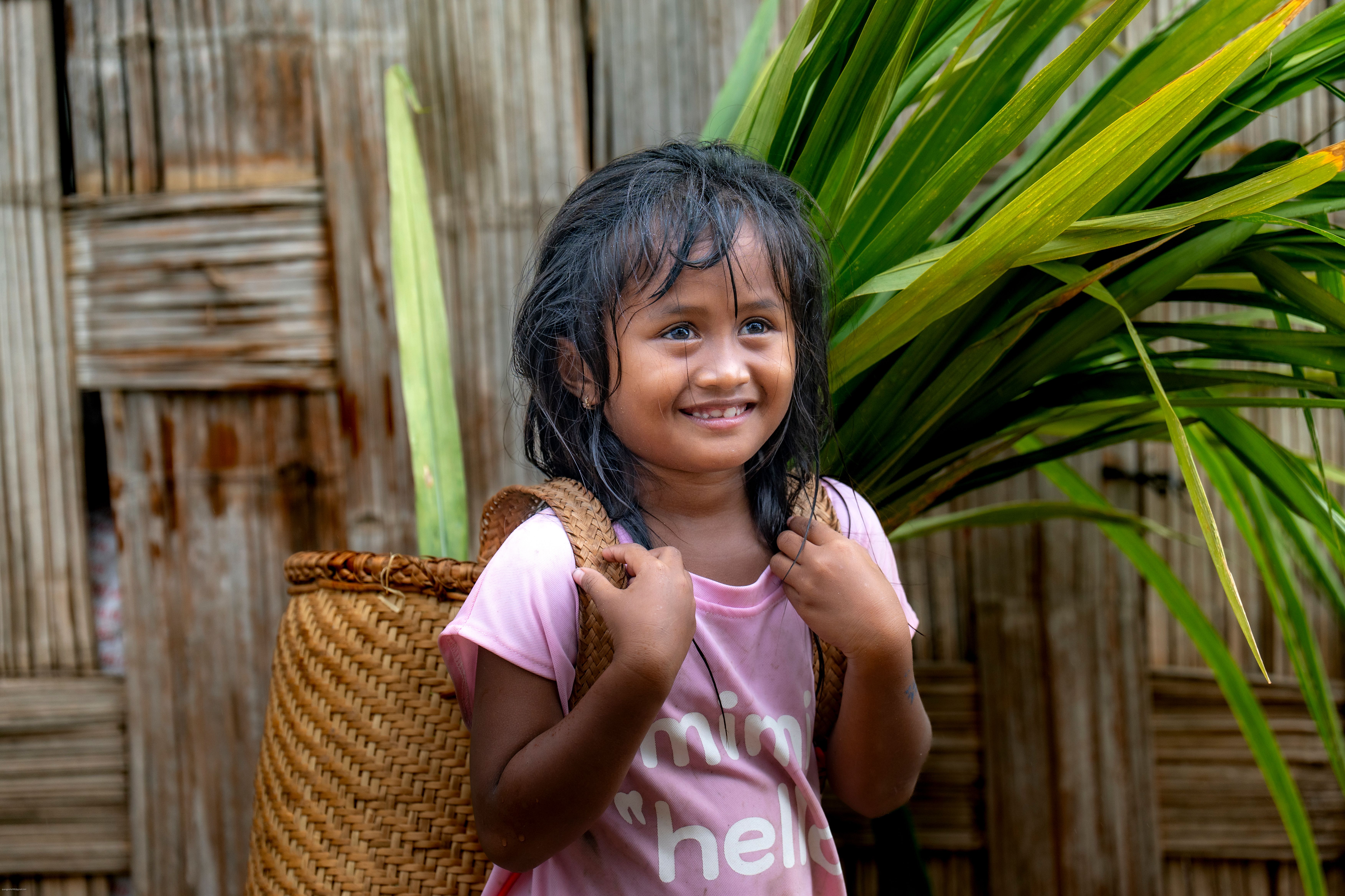 Joyful young girl with a straw backpack, standing in a tropical setting.